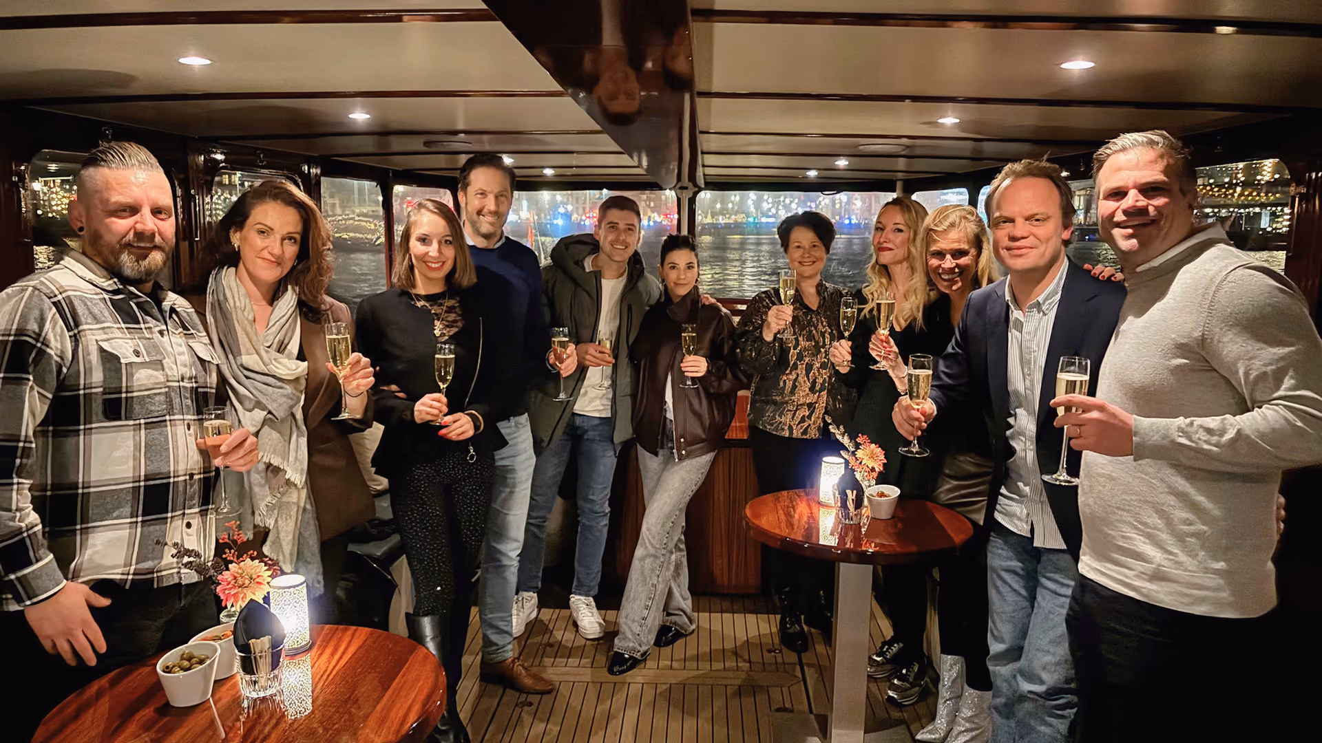 A group of eleven people standing indoors at night, holding champagne glasses, smiling and posing for a photo around wooden tables with small flower arrangements and lit candles.