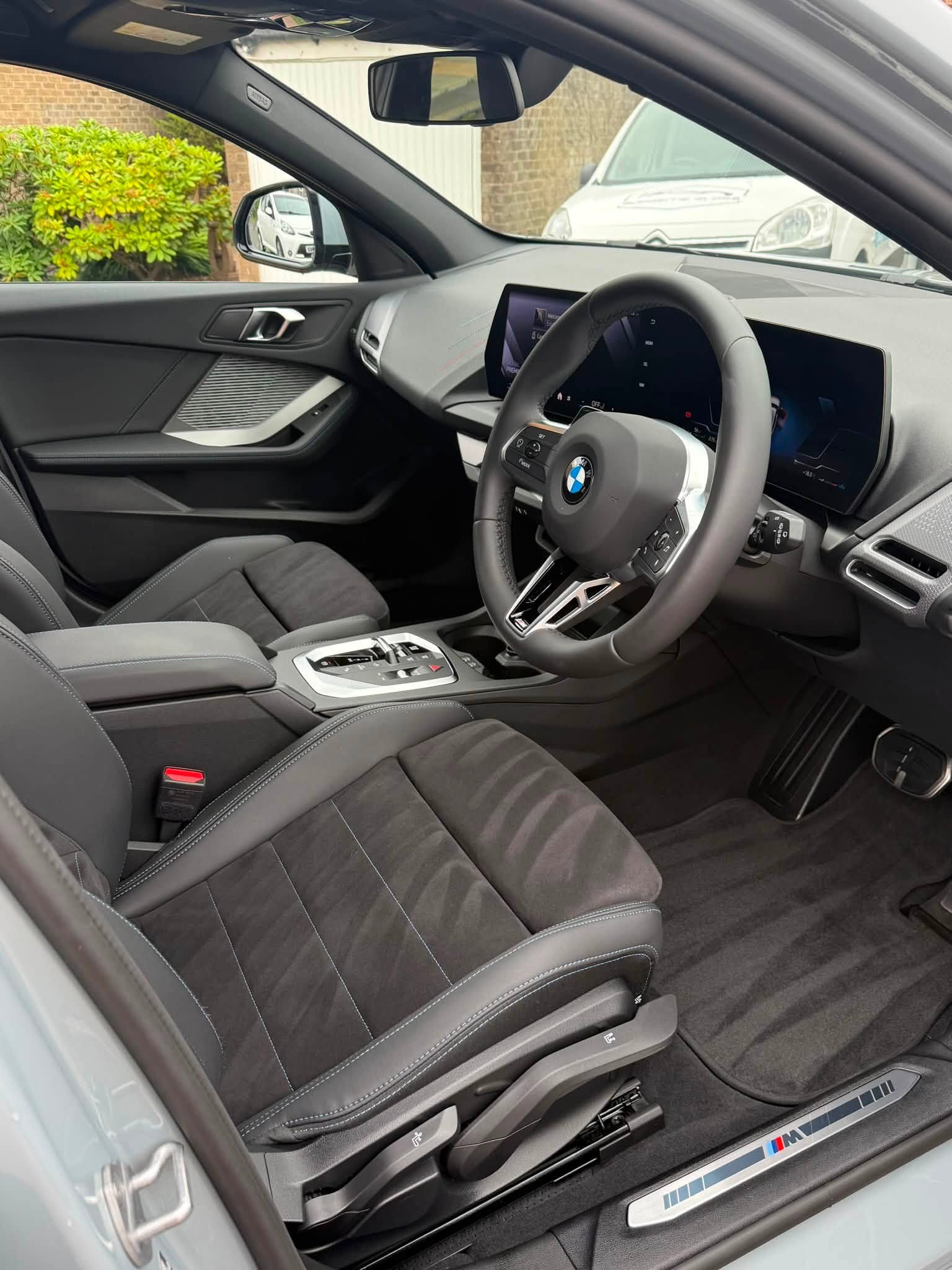 Interior of BMW car showing black suede seats, steering wheel with logo, and dashboard with digital displays.