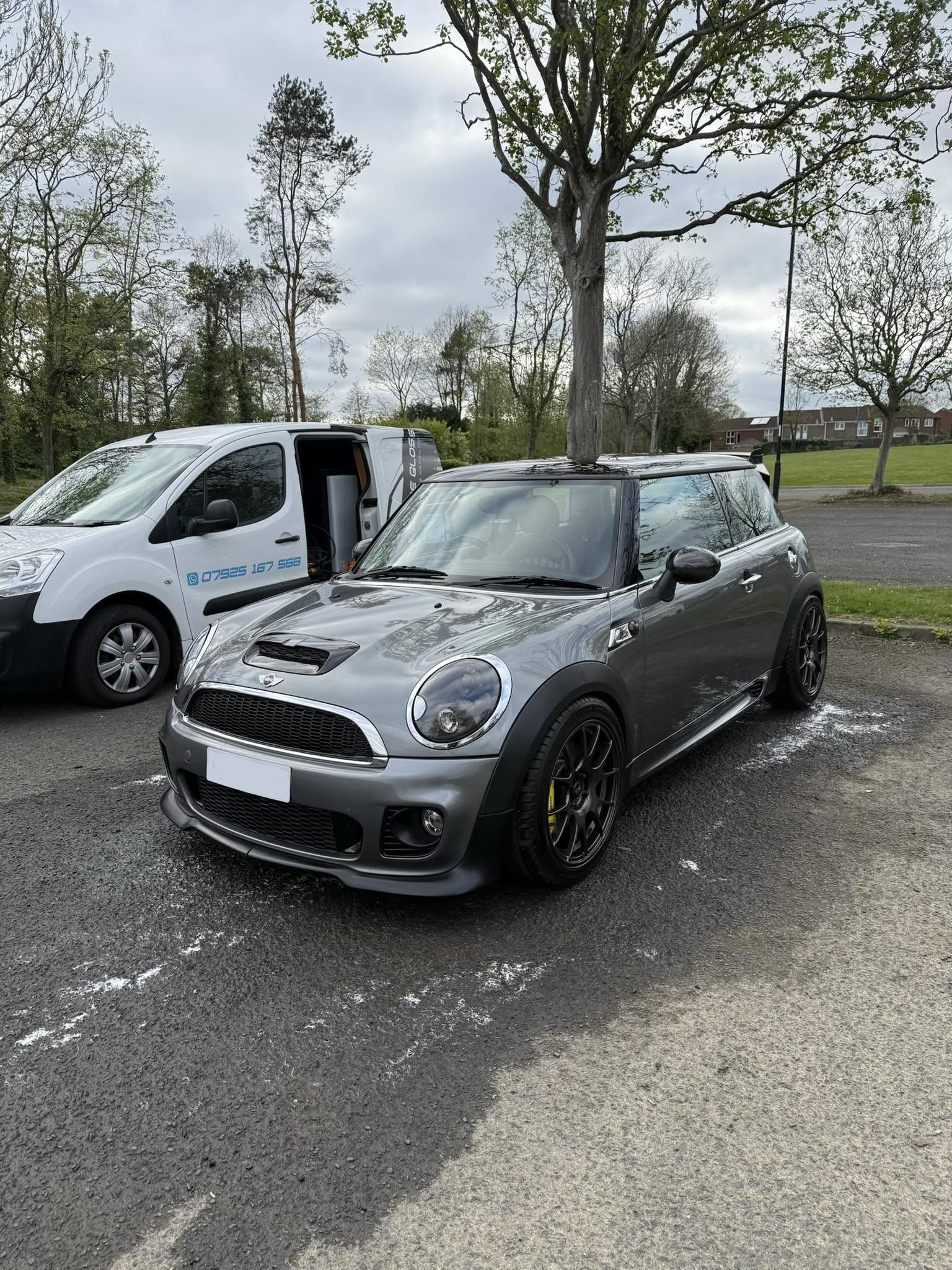 Gray Mini Cooper parked on asphalt near a white van with trees and houses in background.