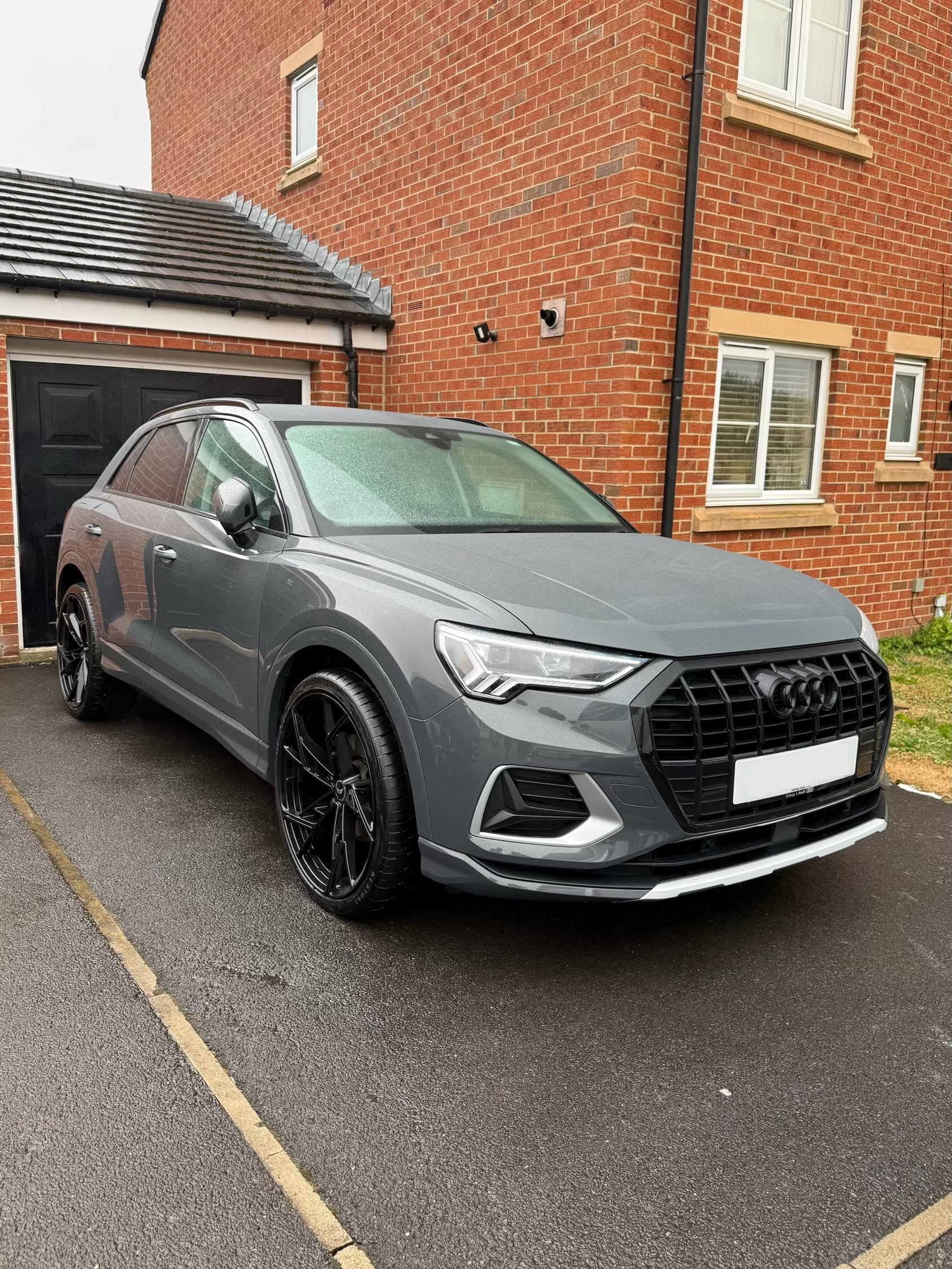 Gray Audi SUV with black rims parked on a driveway in front of a brick house with white windows.