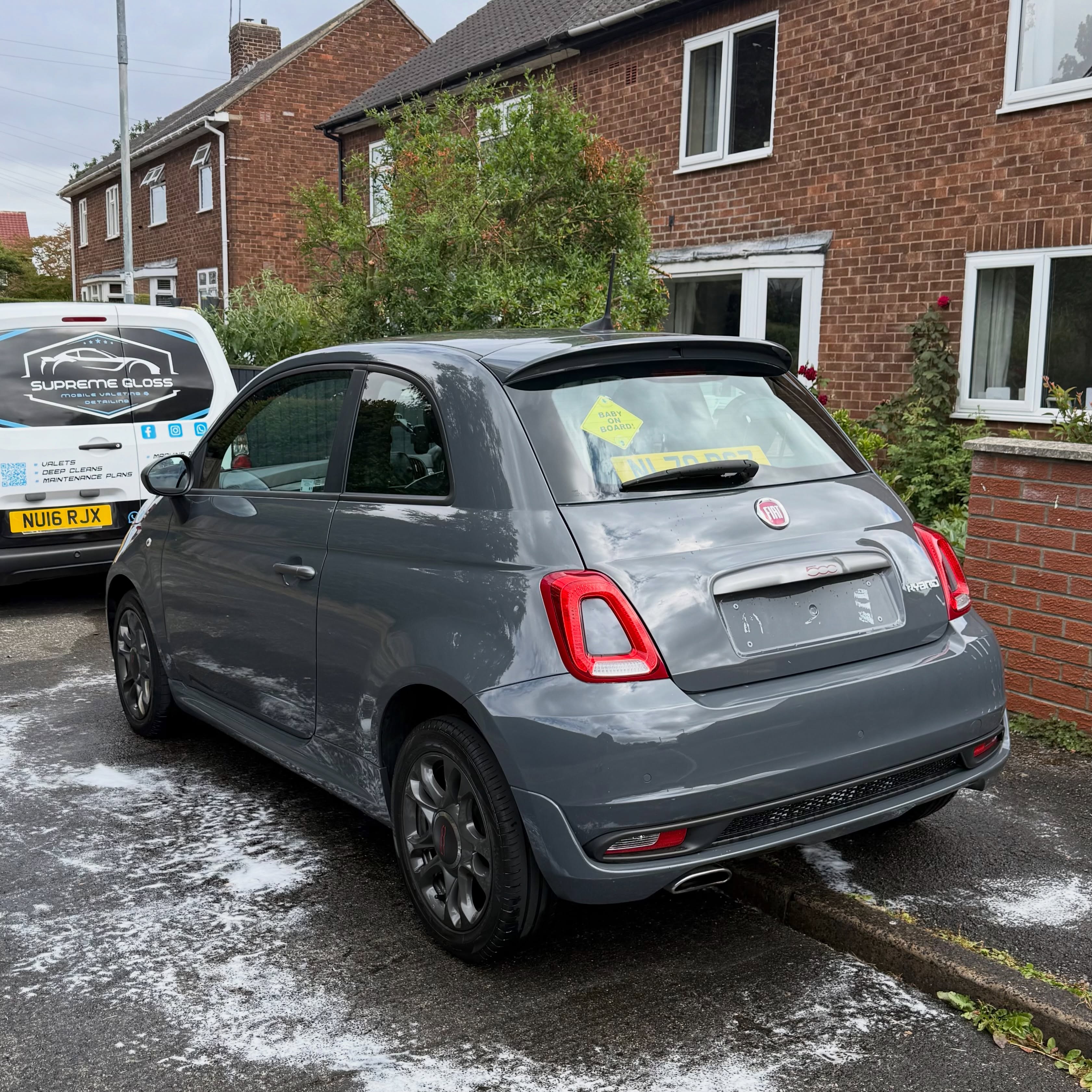 Gray Fiat 500 parked on street near brick houses and a white van with cleaning service logo.