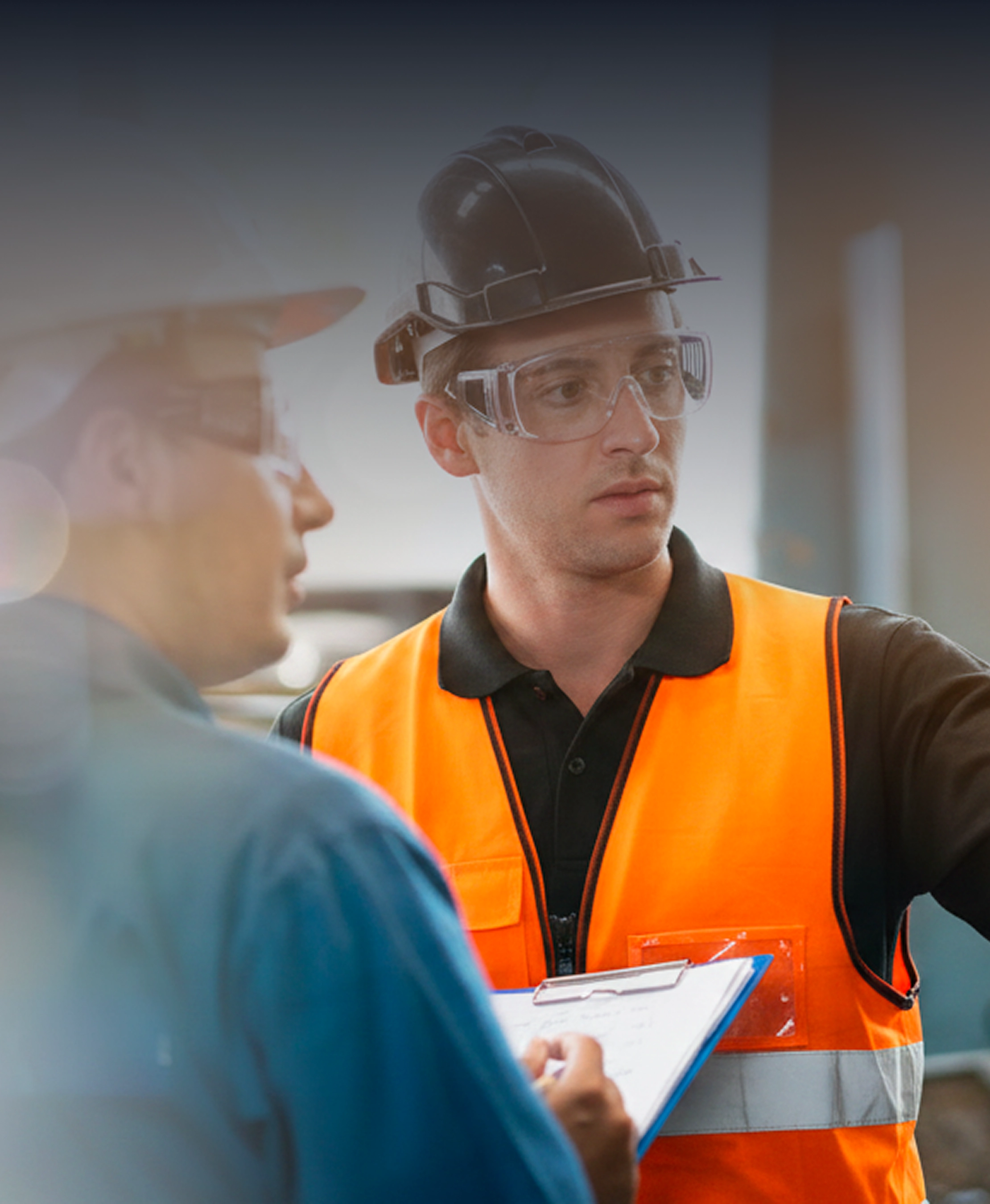 Two industrial workers in safety gear discussing plans, one holds a clipboard wearing an orange vest.