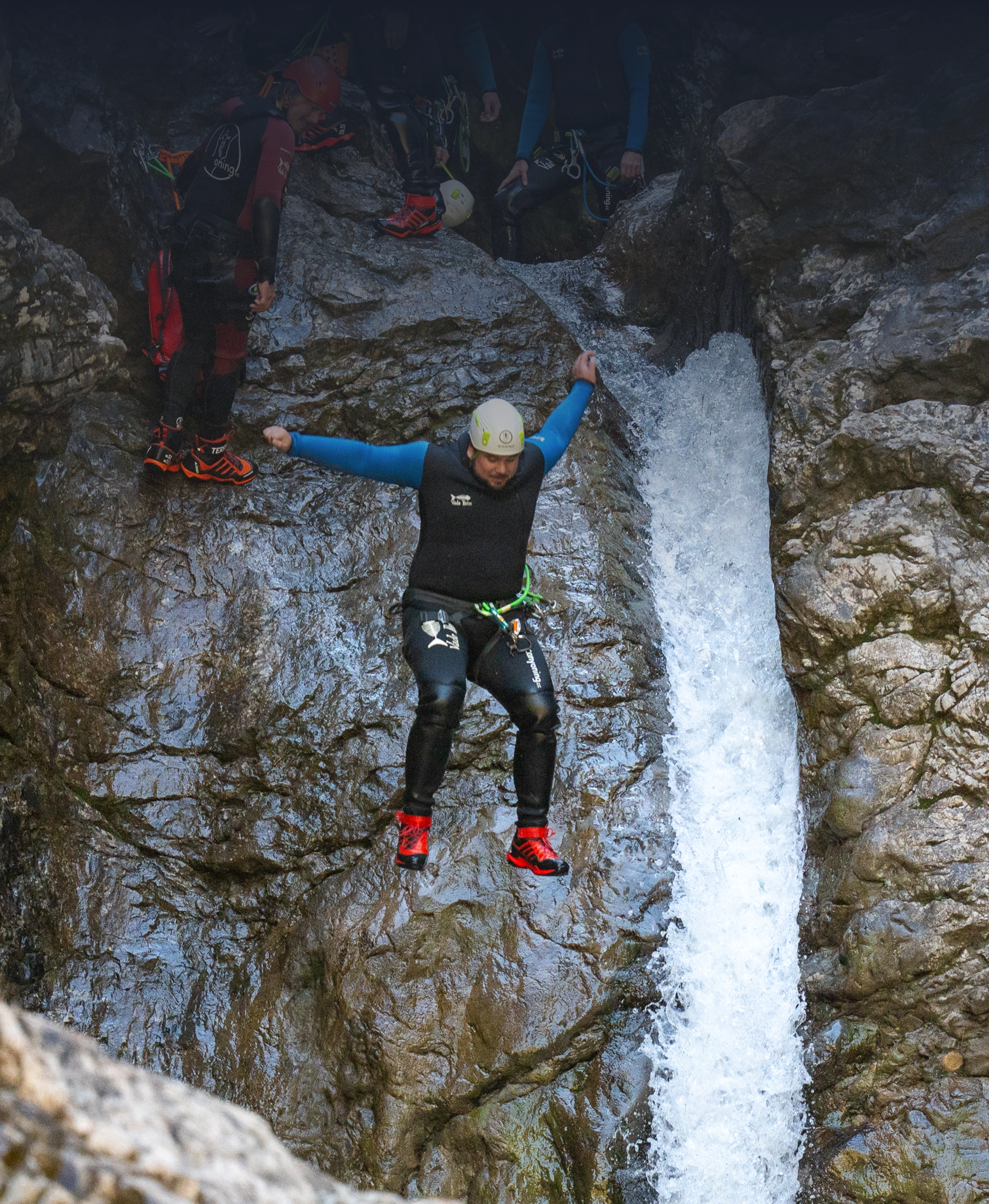 Man in wetsuit and helmet jumping down rocky waterfall during canyoning adventure.