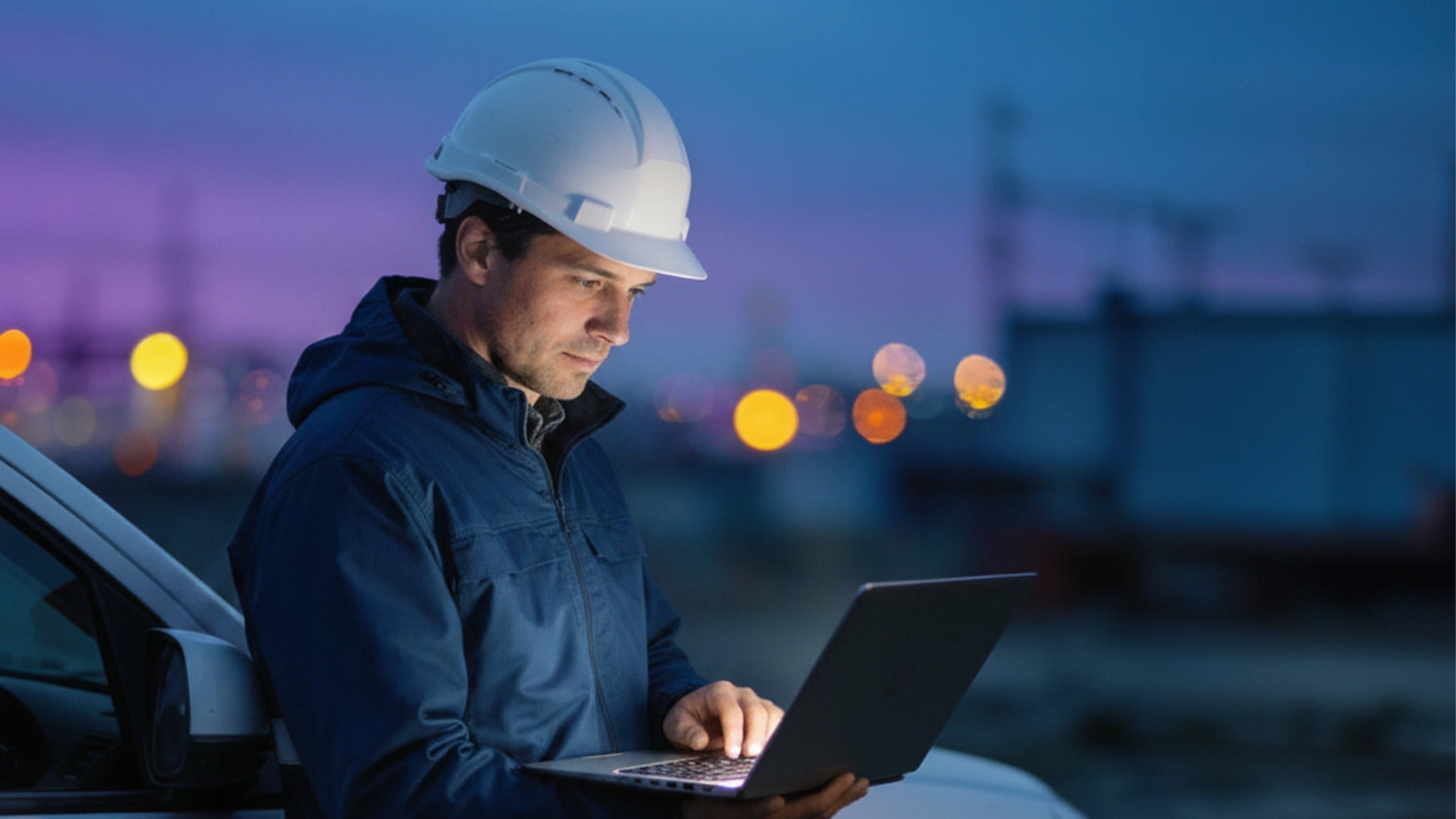 Man wearing white hard hat and blue jacket using laptop outdoors at dusk near vehicle and industrial site.