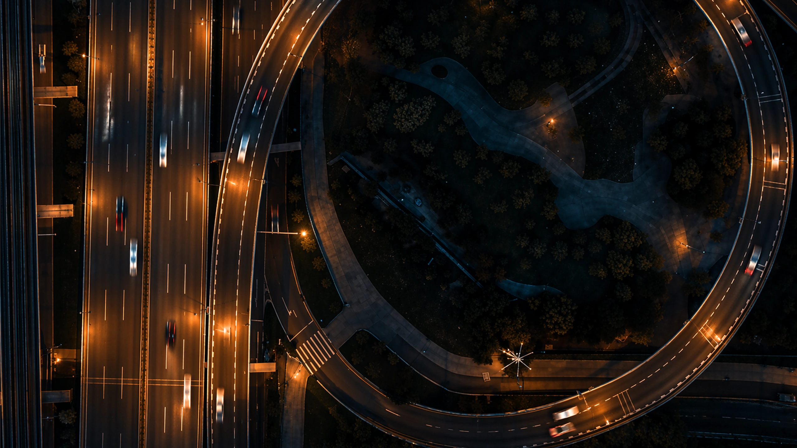 Aerial night view of a lit highway with fast-moving cars and an adjacent curved road with streetlights.
