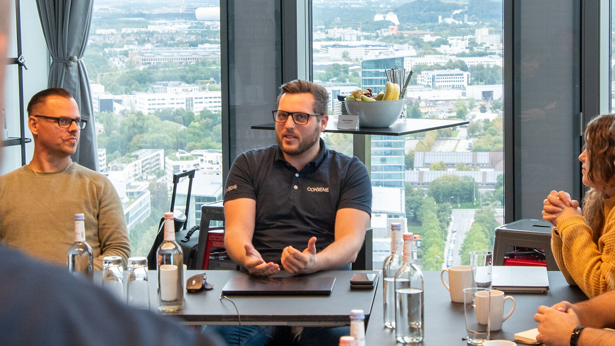 People having a meeting around a table with laptops, water bottles, and a city view behind windows.