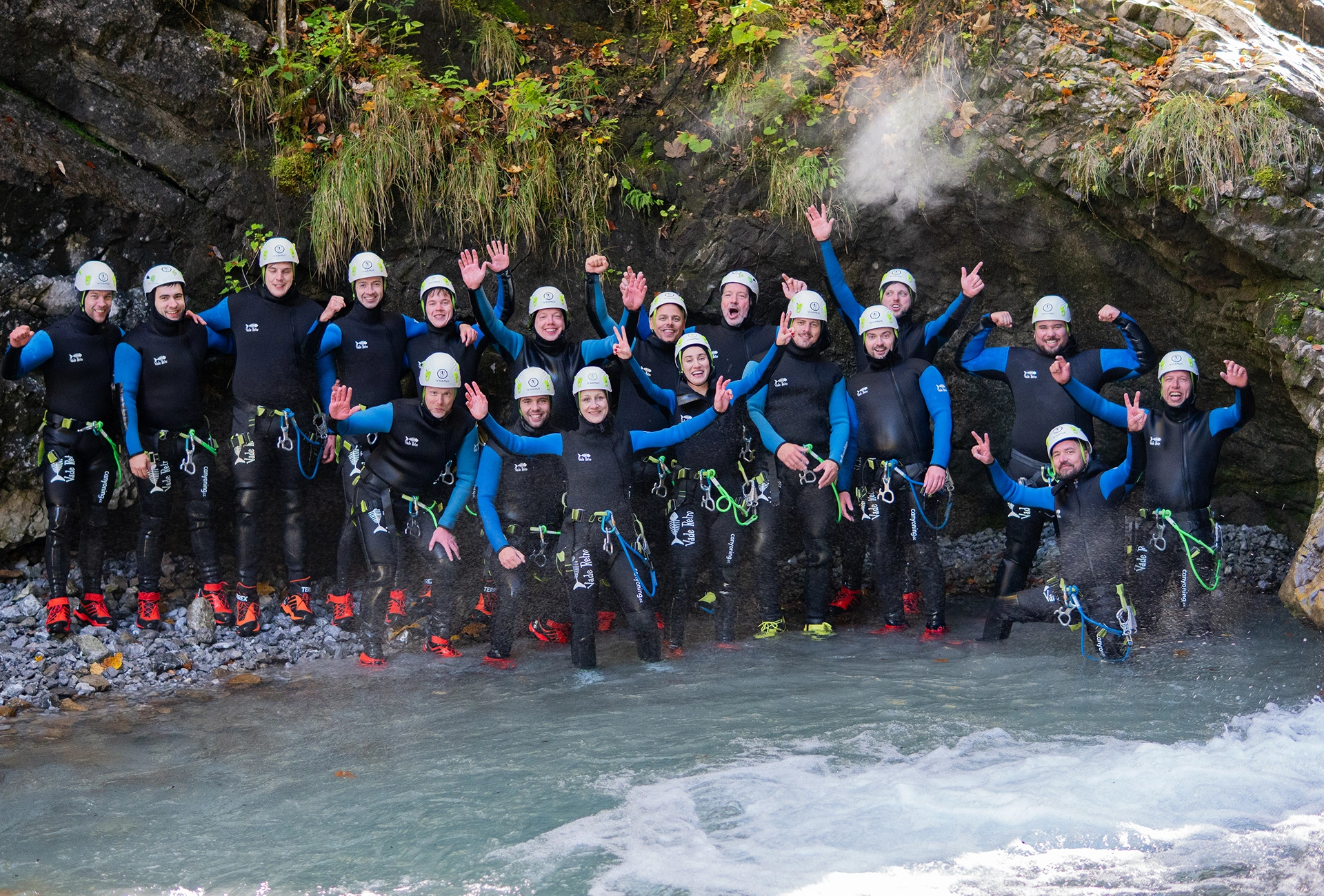 Group of people in wetsuits and helmets standing in water by rocky cave, posing and cheering.