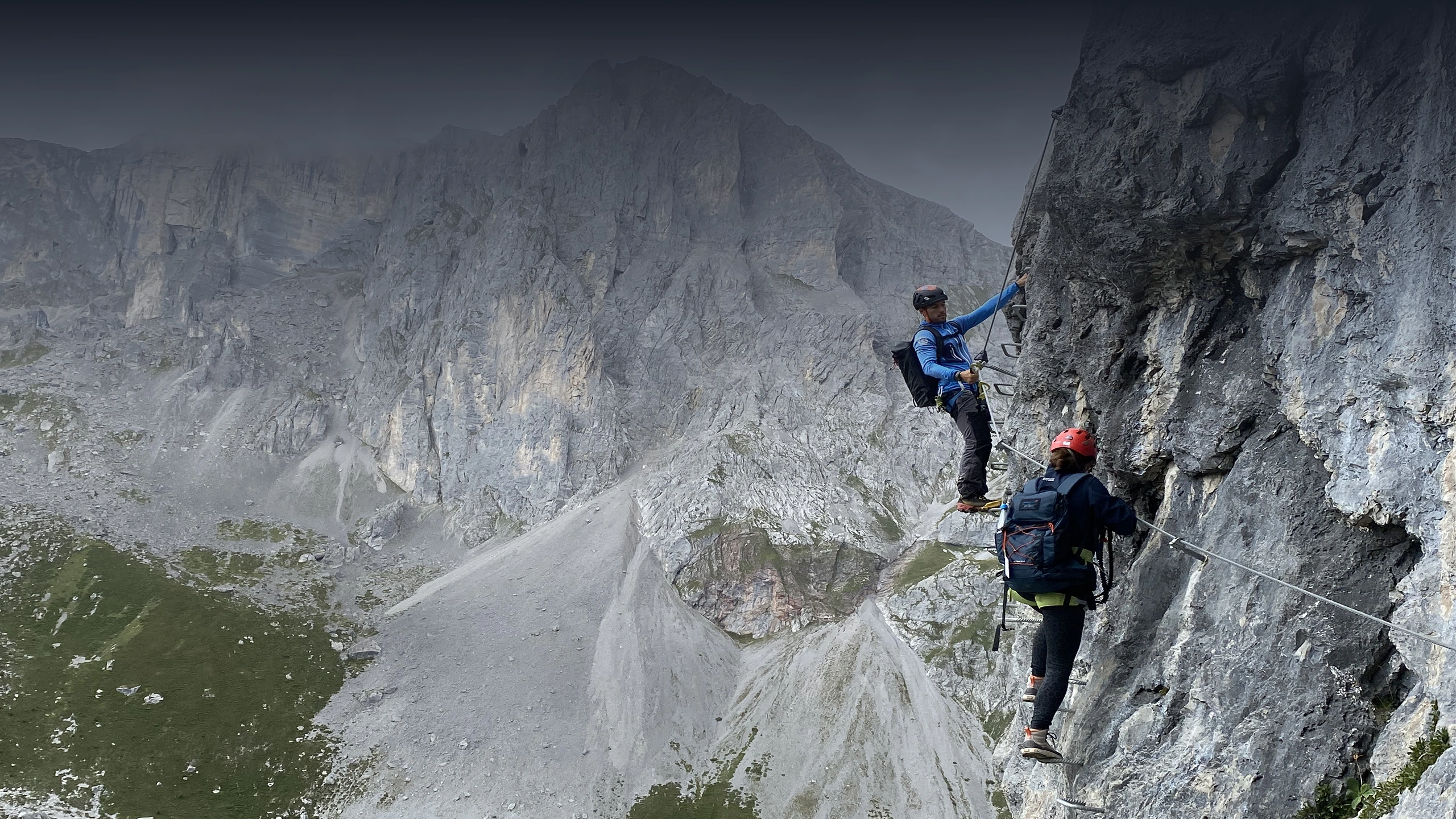 Two climbers with helmets and backpacks ascending a steep rocky mountain face using metal rungs.