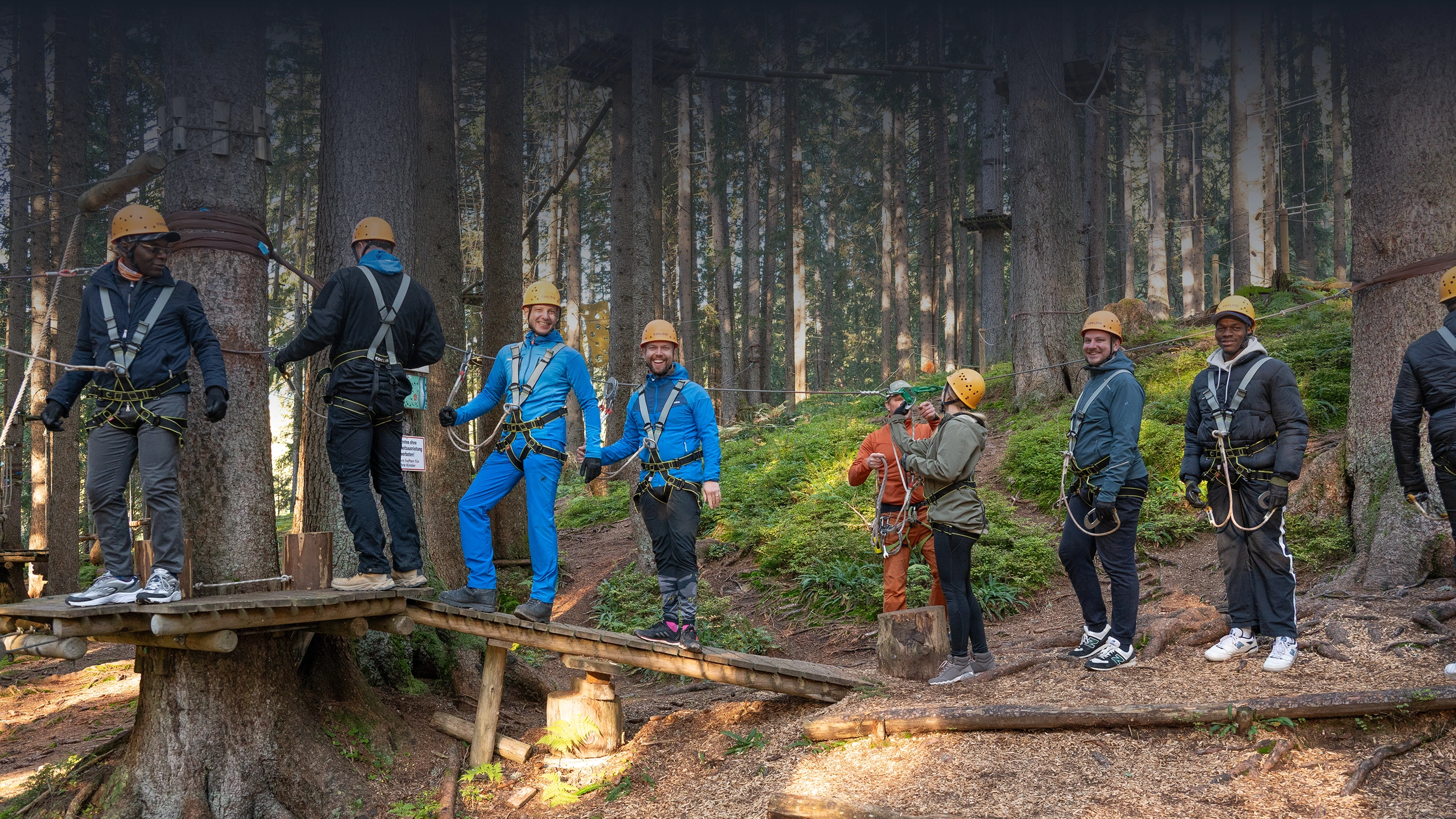 Group of people wearing helmets and harnesses preparing for a forest ropes course adventure.