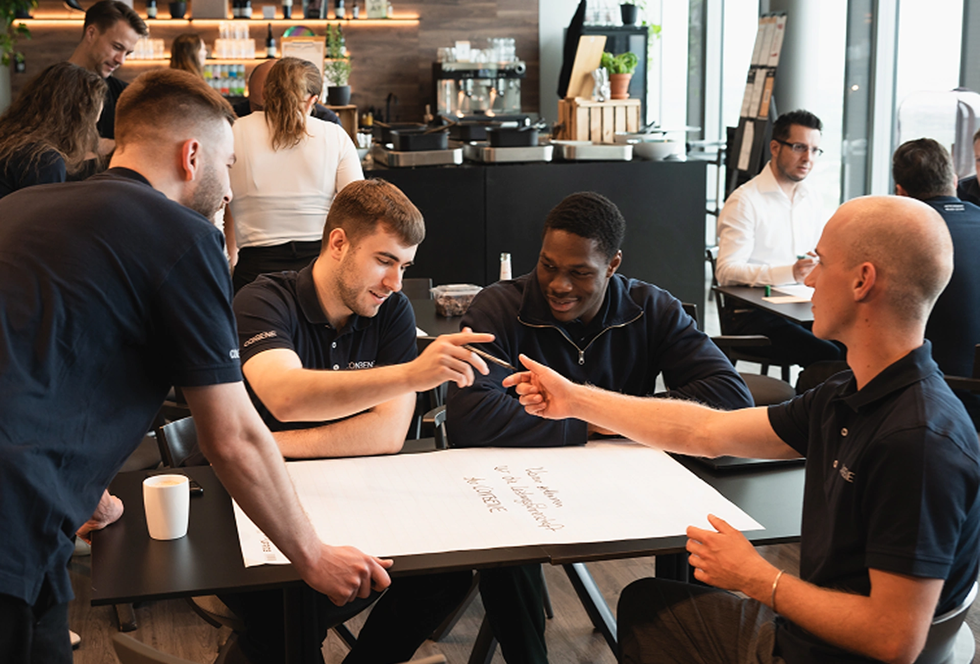 Four men collaborating over a large paper with notes in a modern cafe setting.