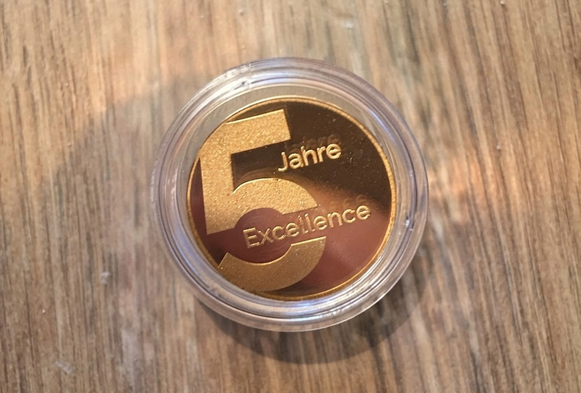 Gold commemorative coin in a clear case reading '5 Jahre Excellence' on a wooden surface.