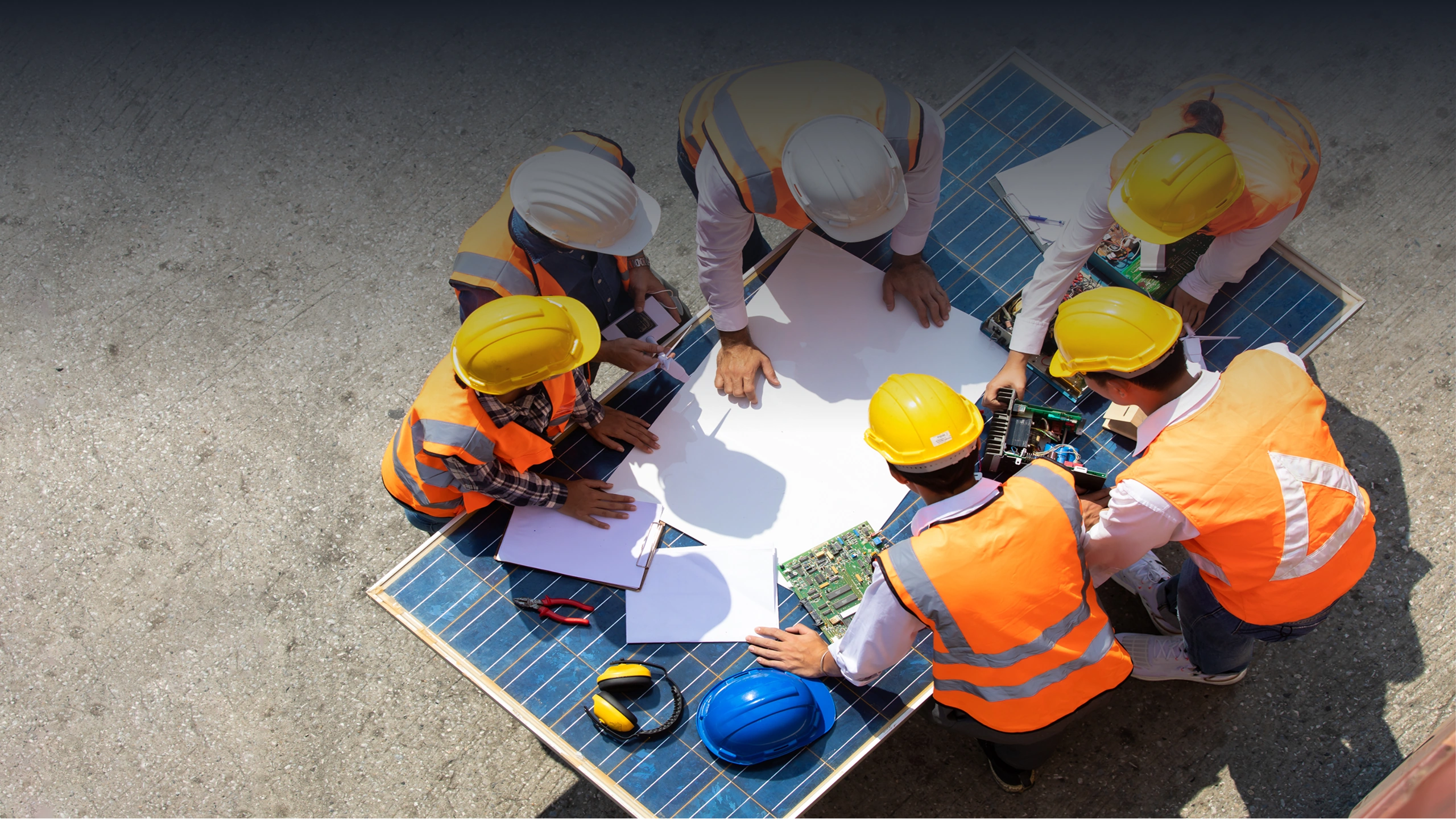 Engineers in hard hats and orange vests reviewing blueprints on a solar panel outdoors.