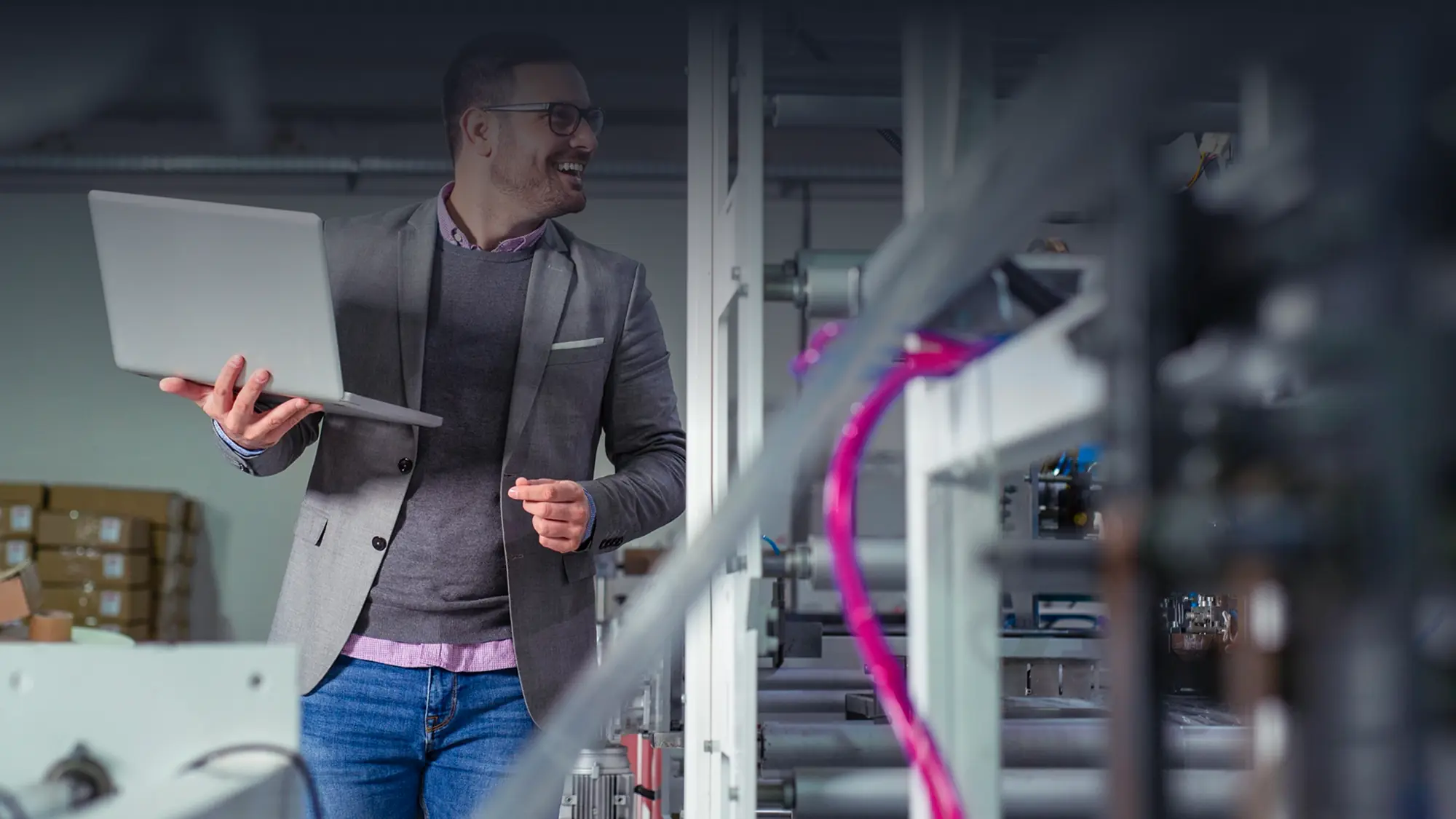 Man in glasses and blazer holding laptop, smiling, standing in a server room.