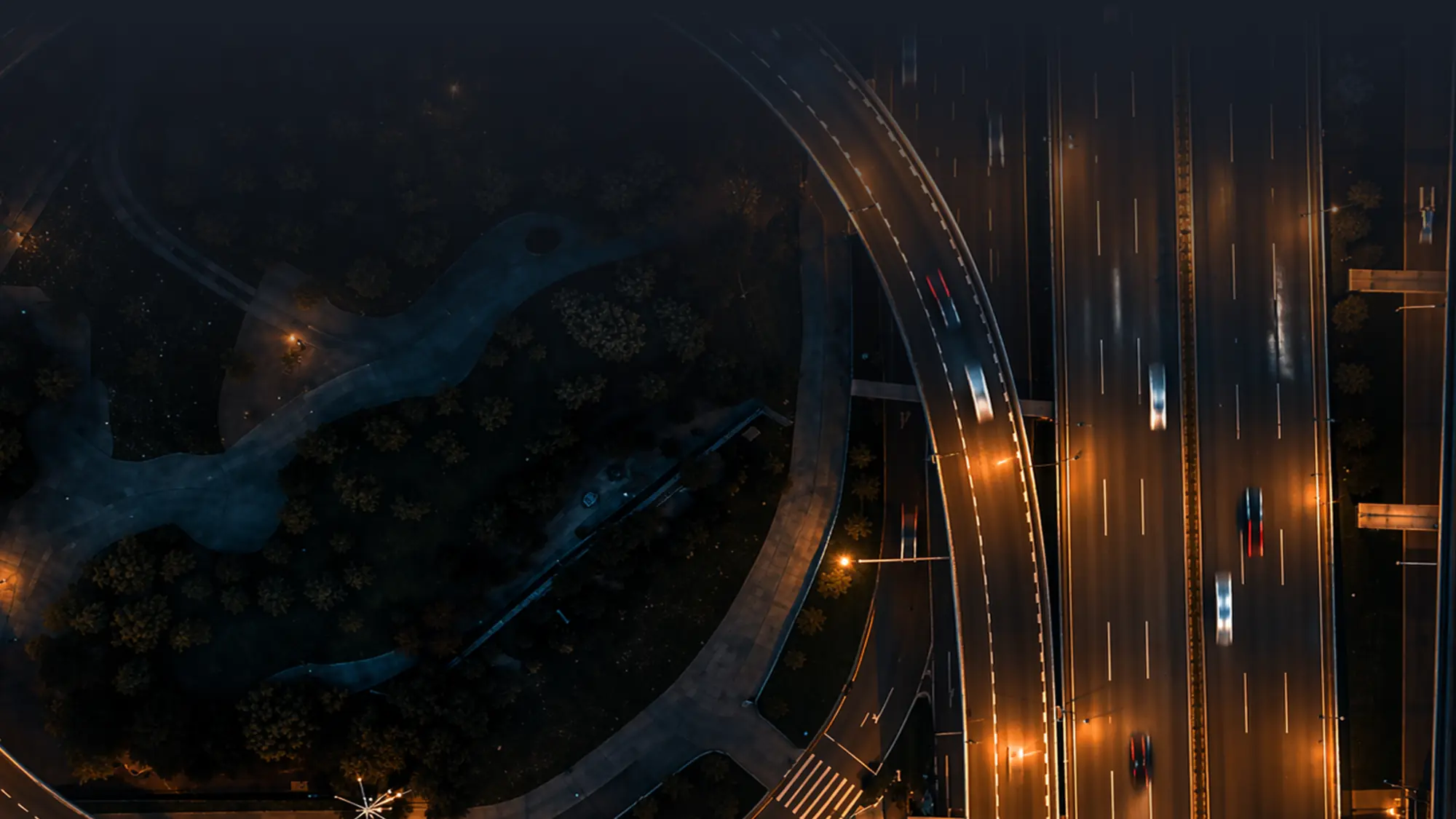 Aerial night view of a lit highway with fast-moving cars and an adjacent curved road with streetlights.