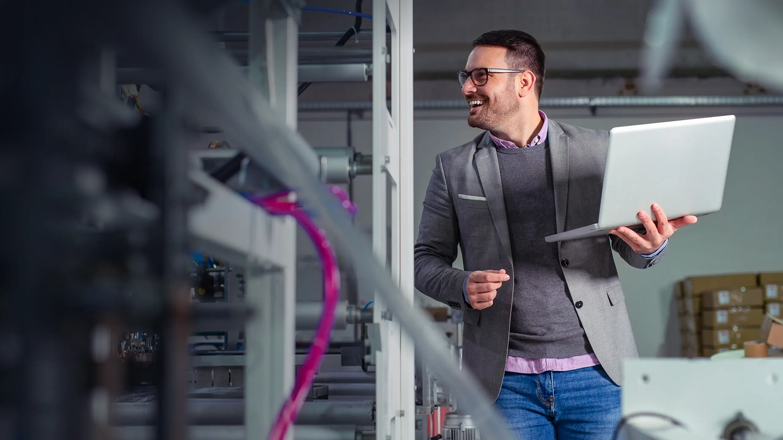 Man in glasses and blazer holding laptop, smiling, standing in a server room.