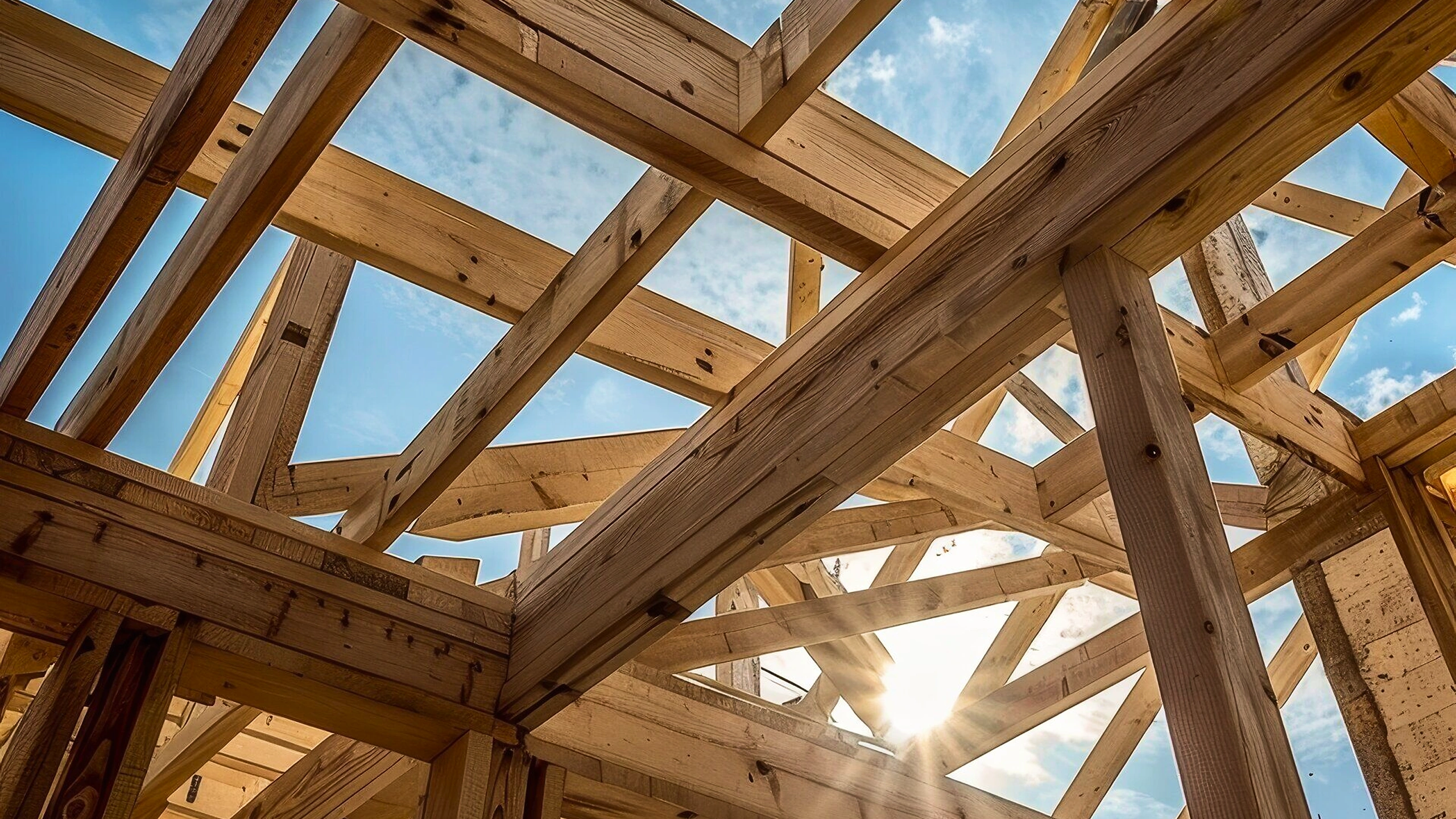 Sunlight shines through the wooden framework of a building under construction against a blue sky.