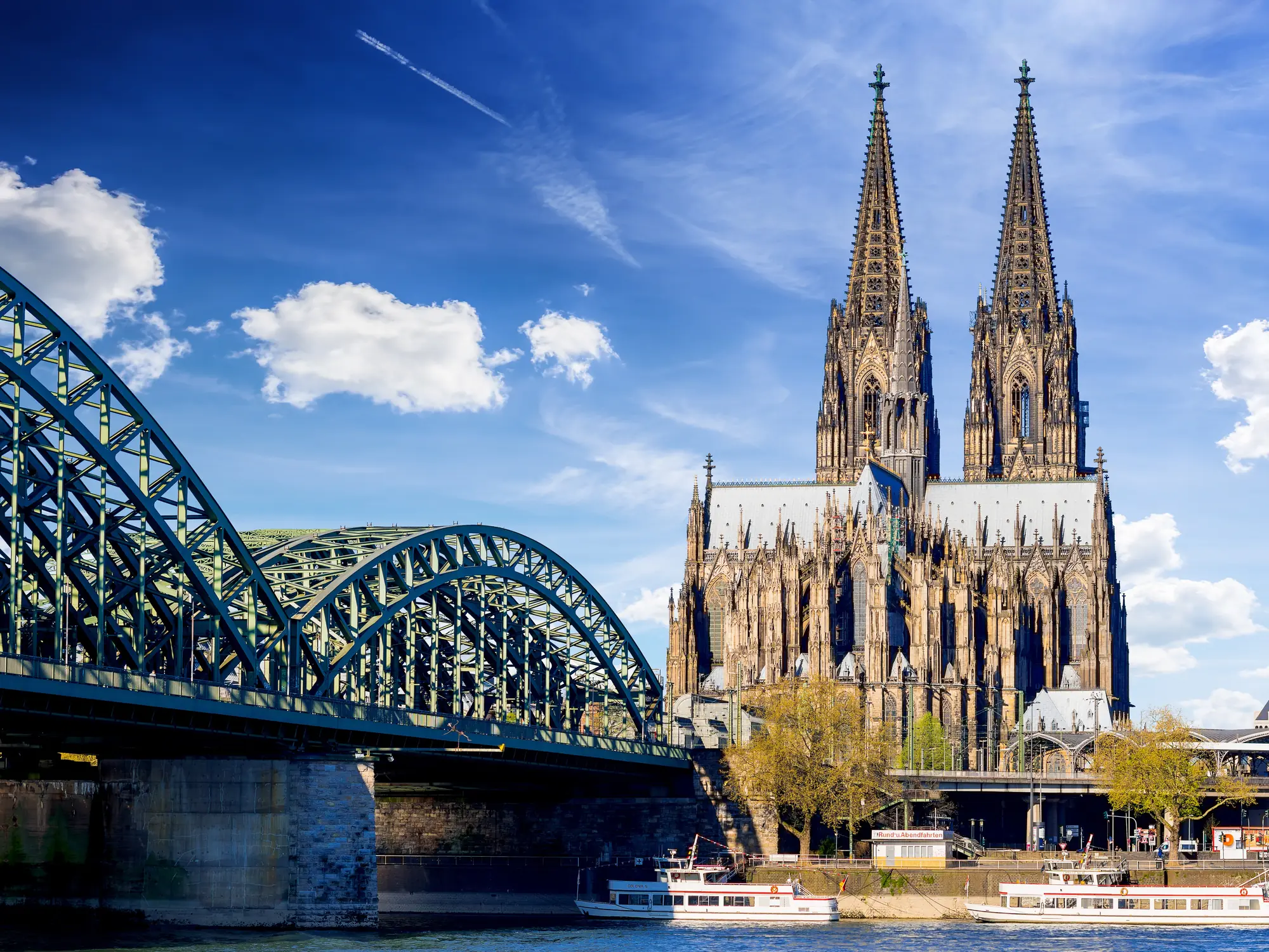 Cologne Cathedral with twin spires next to Hohenzollern Bridge over the Rhine River in sunny weather.