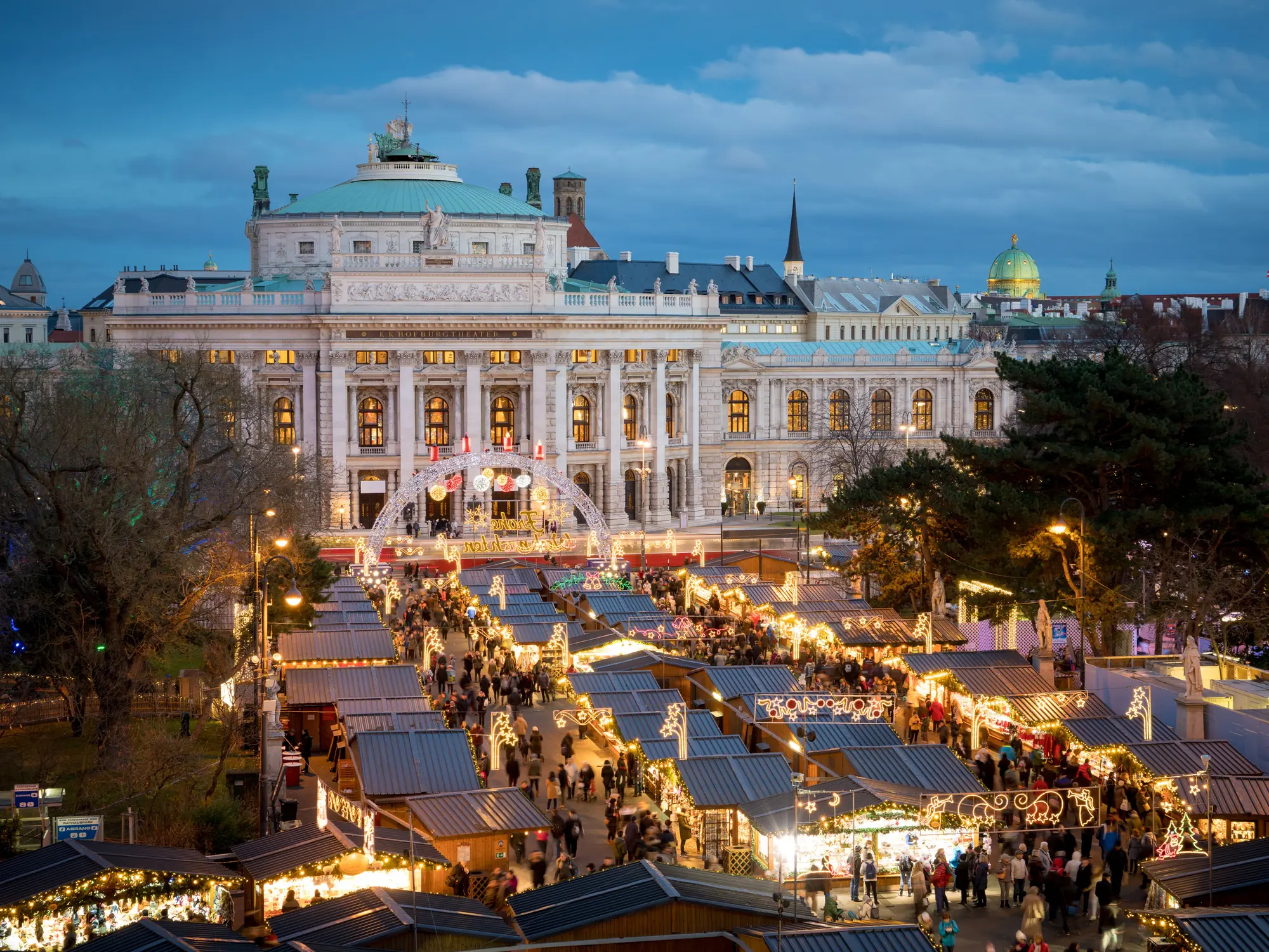 Evening view of illuminated Christmas market stalls with a historic building in the background in Vienna.