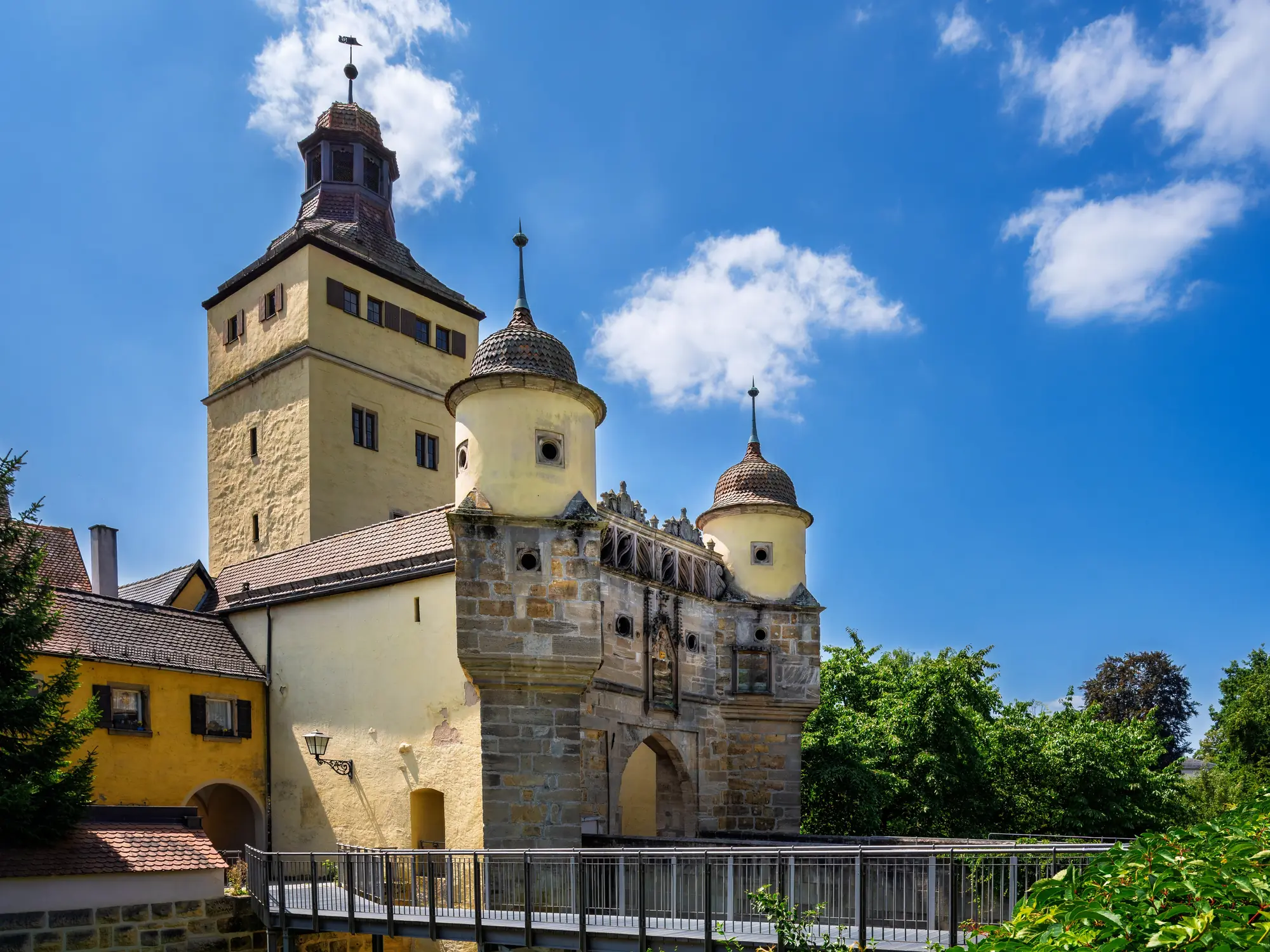 Historic yellow stone gatehouse with towers under blue sky and a railing bridge in front. Ellinger Tor in Weissenburg.