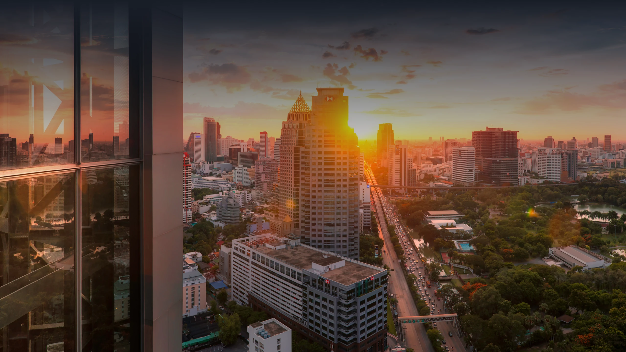 Sunset view of a city skyline with high-rise buildings, a busy road, and green park areas.