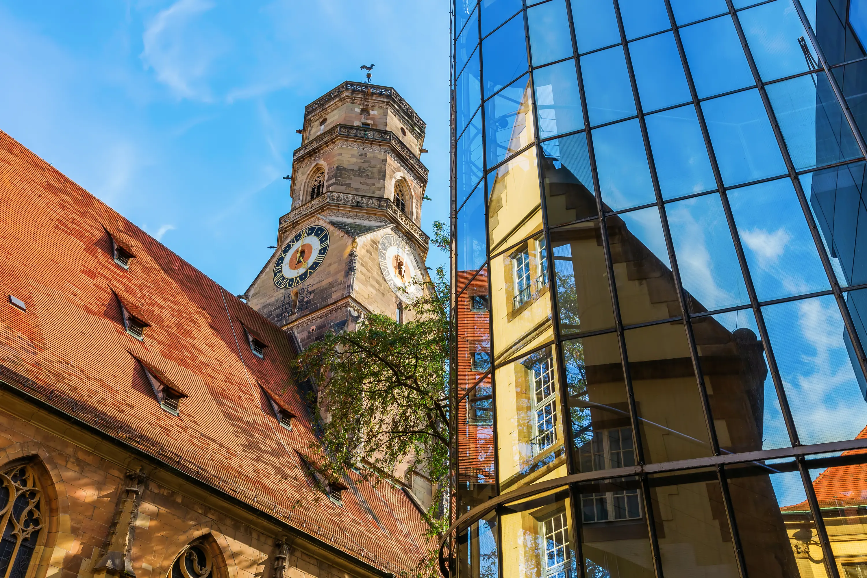 Stone clock tower with a weather vane beside a building with a red tiled roof, reflected in the glass facade of a modern building under a blue sky in Stuttgart