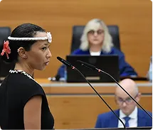 Woman in traditional attire speaking at a courtroom with judge and lawyer in the background.