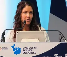 Woman speaking at a podium with One Ocean Science Congress signage and blue-and-white background.