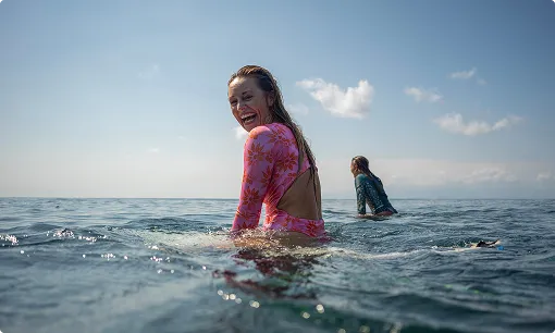 Two surfers sitting on boards in the ocean, woman in pink smiling at camera under blue sky.