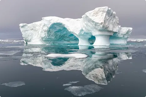 Large iceberg with arches reflecting on calm, icy Arctic water under a gray sky.