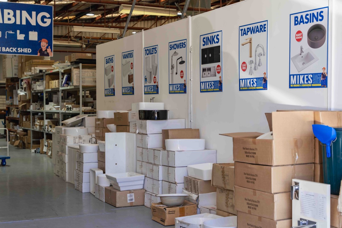 Warehouse interior with stacks of boxed bathroom fixtures such as sinks, basins, toilets, showers, tapware, baths, and vanities, displayed under matching product category signs on a white wall.