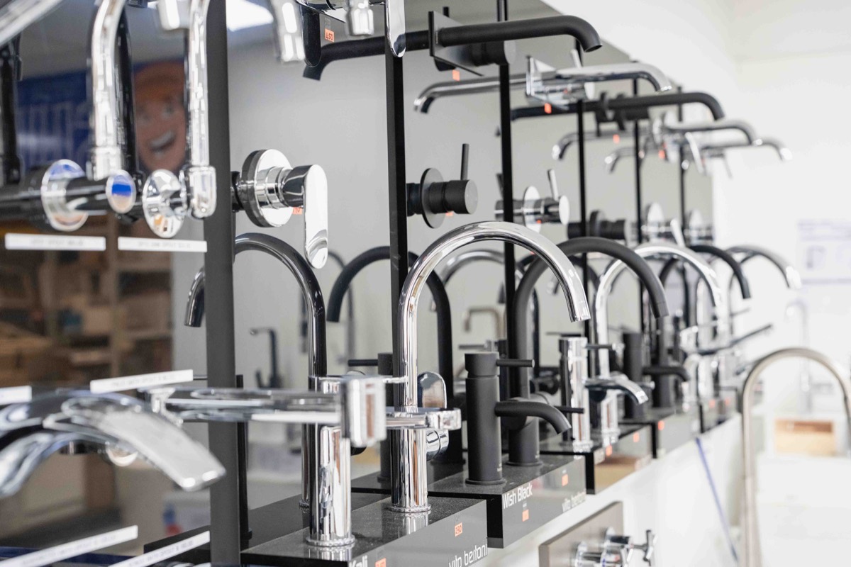 Display of various modern kitchen faucets in chrome and matte black finishes arranged in rows on a showroom counter.