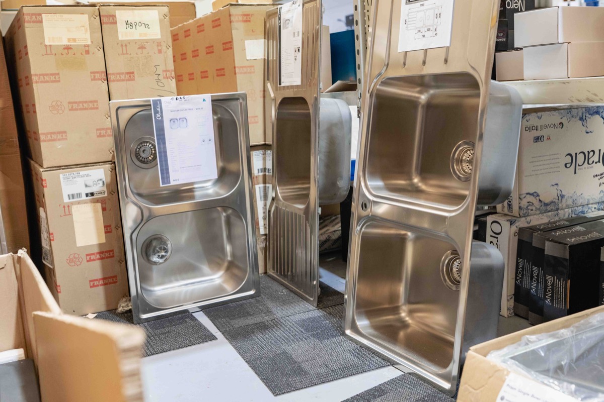 Several stainless steel kitchen sinks with double basins displayed upright on the floor, surrounded by cardboard boxes in a storage or warehouse setting.
