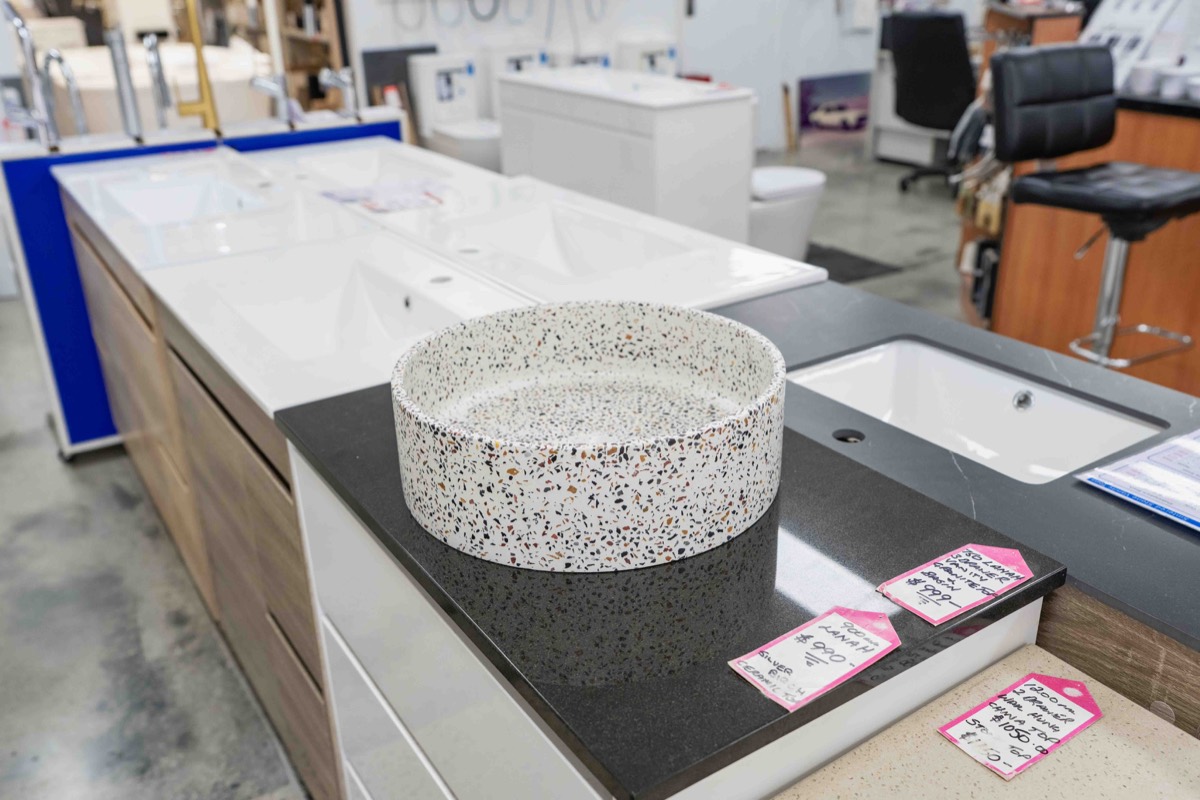 Display of various bathroom sink countertops and basins in a showroom, including a speckled round vessel sink on a black countertop and rectangular undermount sinks on white and beige countertops.