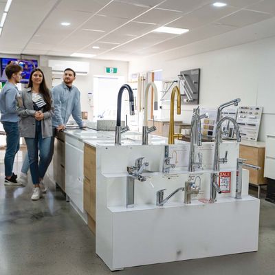 Interior of a showroom displaying various modern kitchen and bathroom faucets mounted on white platforms, with three people walking and observing the products in the background.
