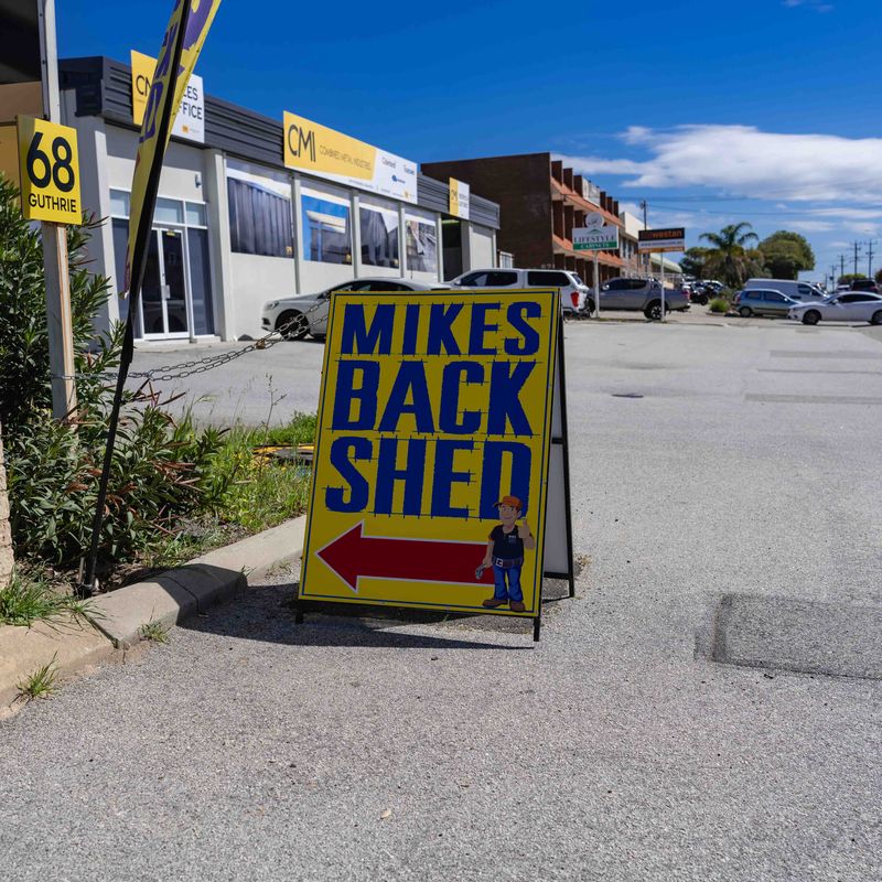 Yellow and blue sign on a sidewalk with the text "MIKES BACK SHED" and a left-pointing red arrow, next to a small cartoon figure of a man in a cap and overalls, directing to a location behind a building with parked cars and business signs.