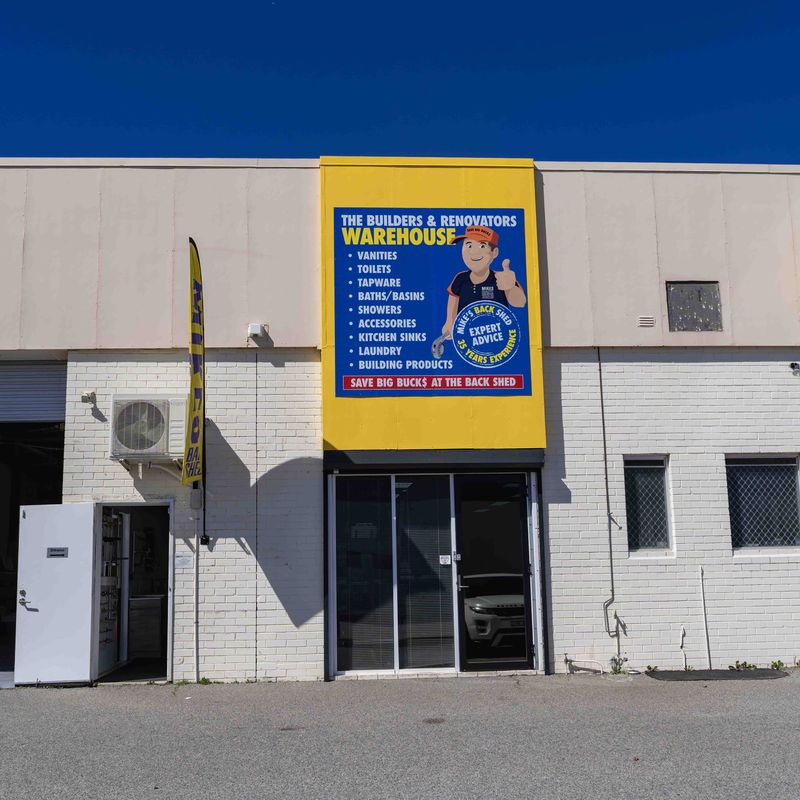 Front view of a white brick warehouse building with a yellow sign above a glass door listing products such as vanities, toilets, tapware, baths/basins, showers, accessories, kitchen sinks, laundry, and building products with a cartoon character and text 'The Builders & Renovators Warehouse' and 'Save Big Bucks at the Back Shed.'