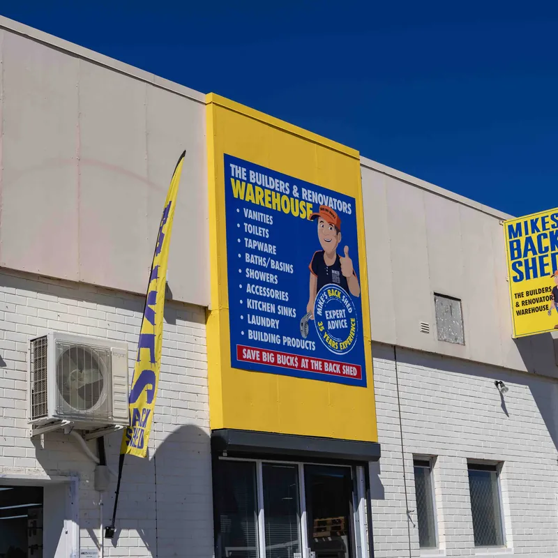 Exterior wall of a building with a large blue and yellow sign listing products including vanities, toilets, tapware, baths, showers, accessories, kitchen sinks, laundry, and building products. The sign features a cartoon man giving a thumbs up and the text 'Expert Advice' and 'Save Big Bucks at the Back Shed'. A yellow vertical flag next to the sign reads 'BACK SHED'.
