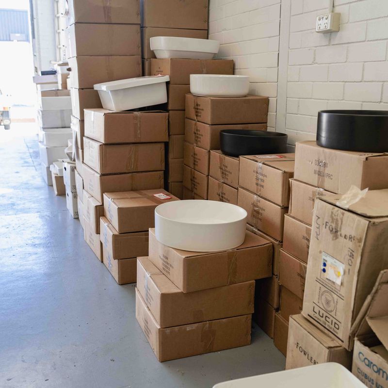 Stacks of cardboard boxes arranged in rows along a warehouse wall, with white and black ceramic basins placed on top of some boxes.