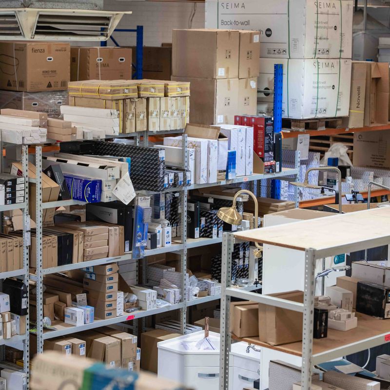 Warehouse shelves stacked with various boxes and packages of different sizes, including plumbing and hardware items, with some visible brand names such as SEIMA and Fienza, and some brass fittings displayed on the shelves.