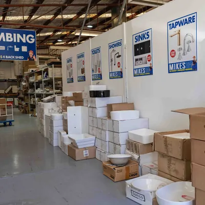 Warehouse aisle with plumbing supplies stacked against a wall under signs labeled Bathrooms, Vanities, Toilets, Showers, Sinks, and Tapware, each with images and the Mikes logo.