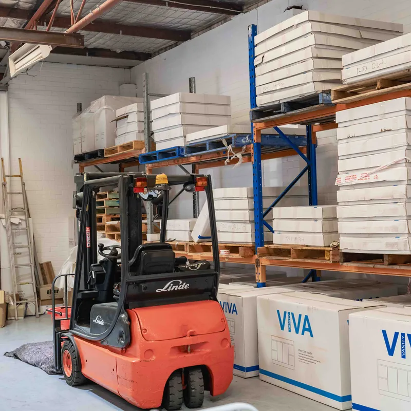 Orange and black Linde forklift parked inside a warehouse with racks holding stacked white boxes and pallets, alongside several large boxed items labeled VIVA.