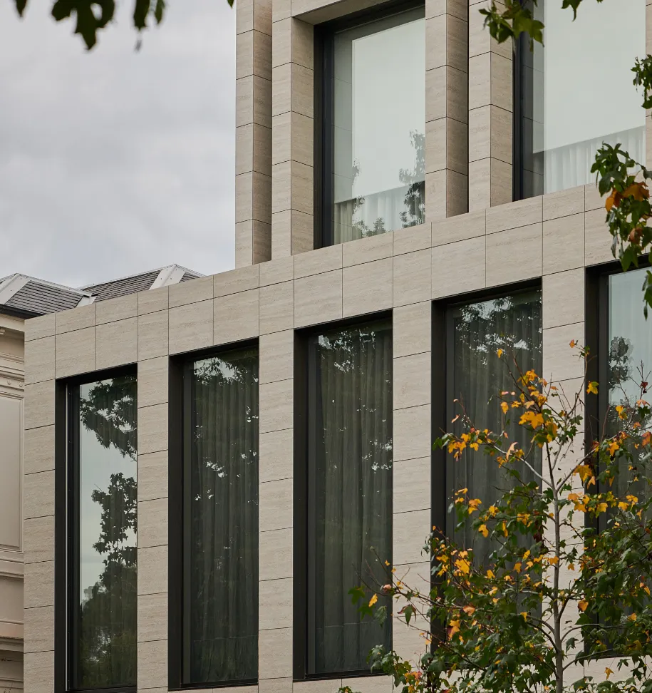 Modern beige stone building facade with large dark-framed windows and green-leafed tree in front.
