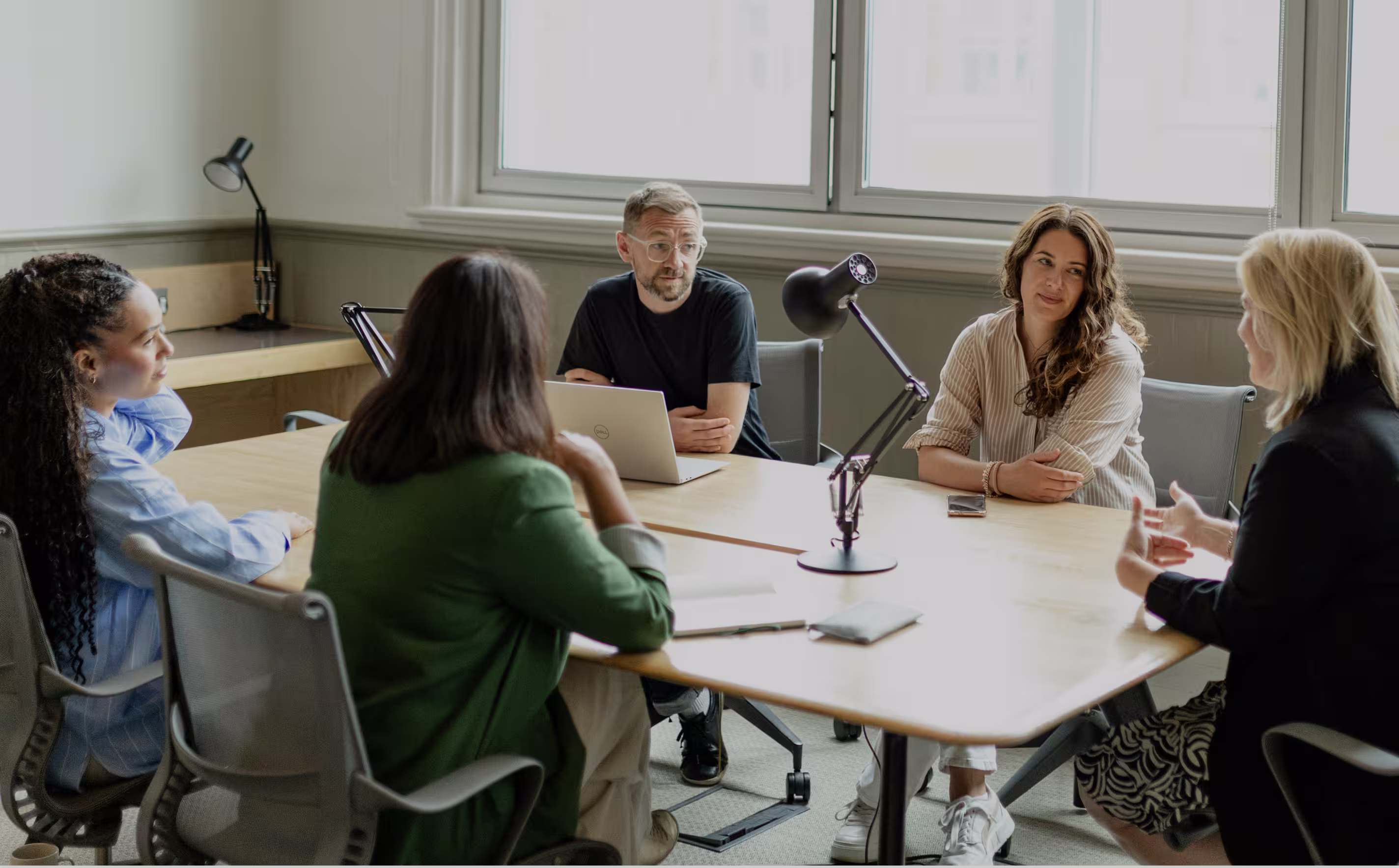 Five diverse professionals in a meeting around a wooden table in a bright office with large windows.
