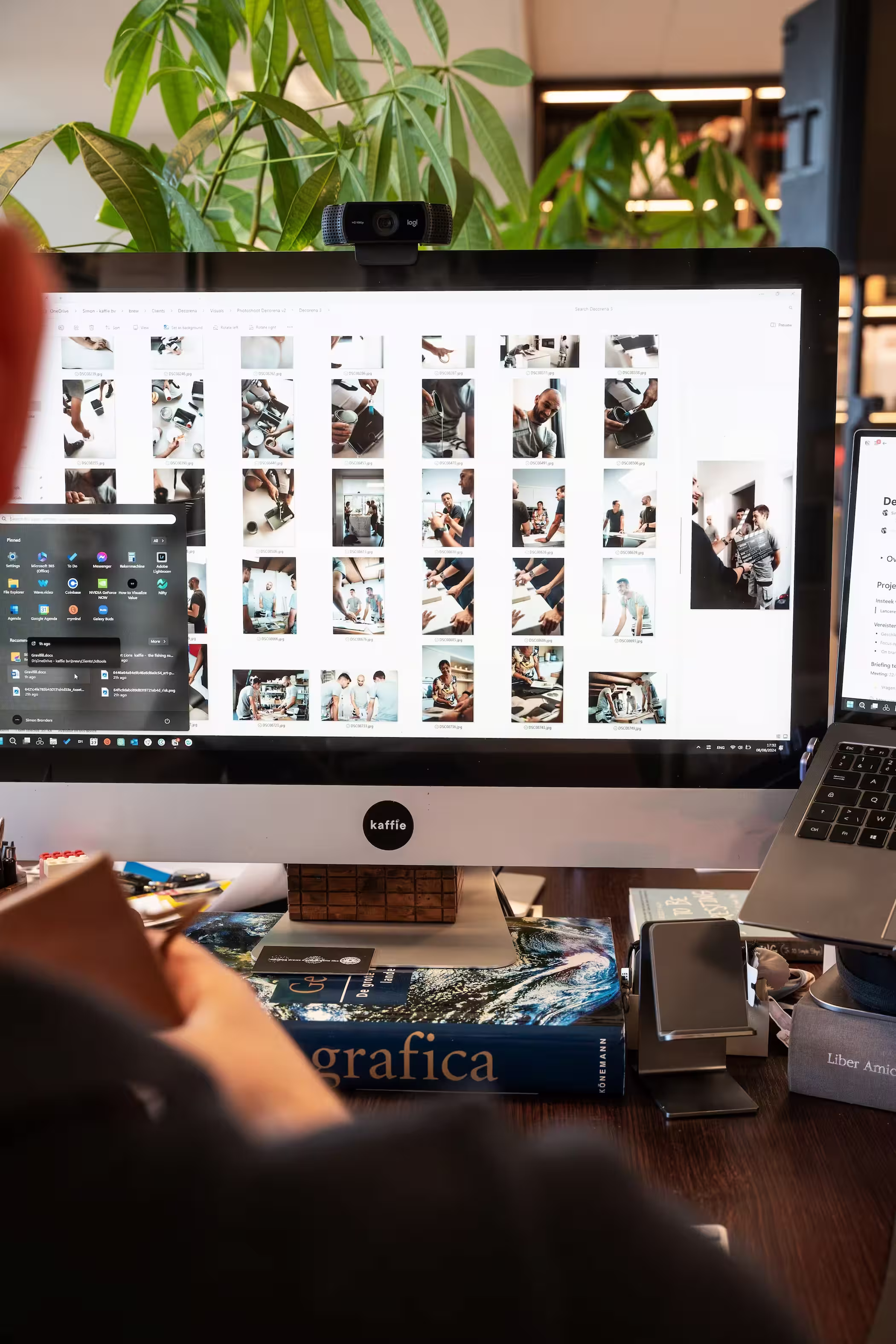 Computer screen displays multiple photo thumbnails on a cluttered desk with plants in the background.