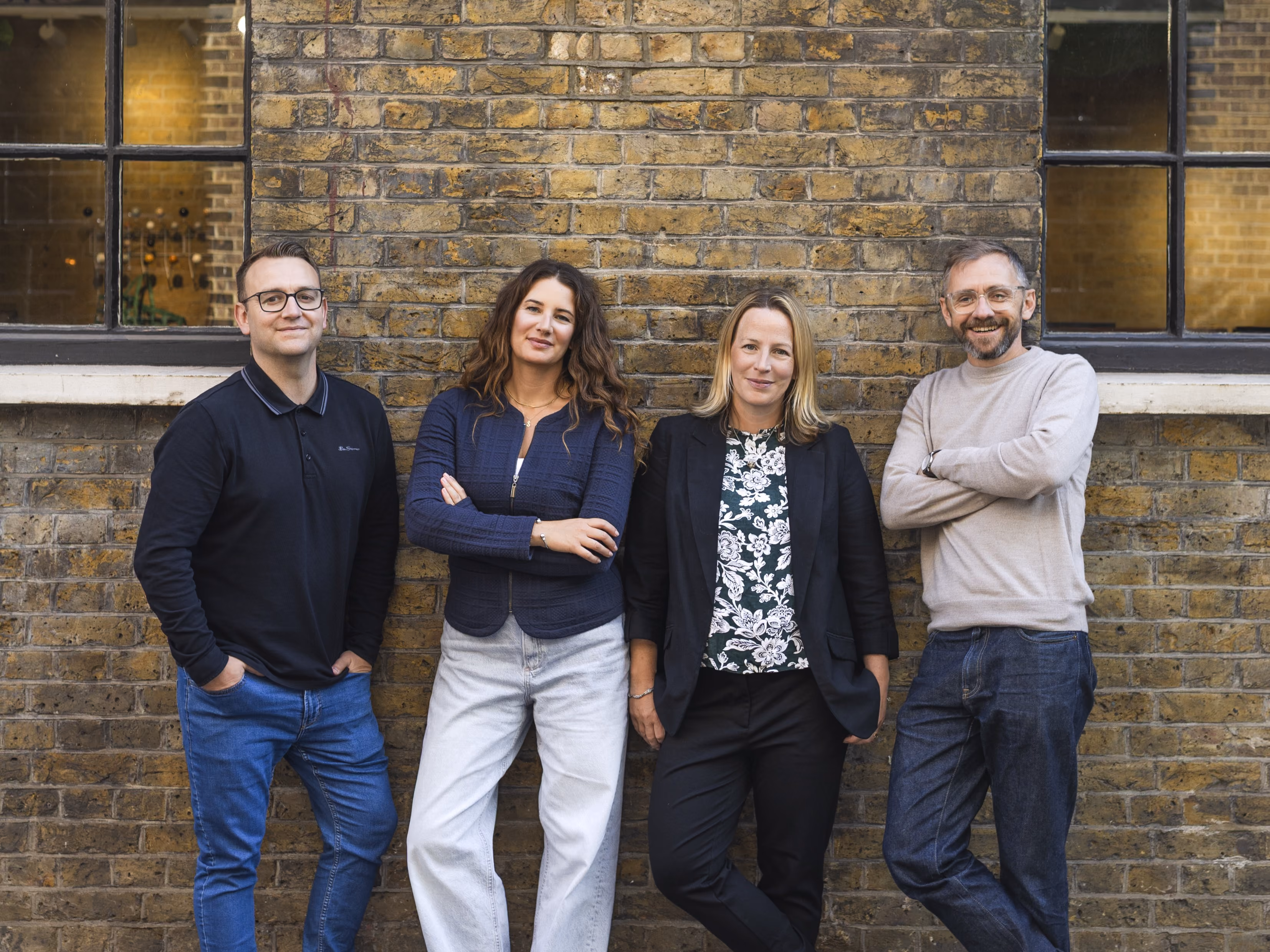 Four adults standing and leaning against a brick wall with windows in the background, two men and two women dressed casually, smiling and posing relaxed.