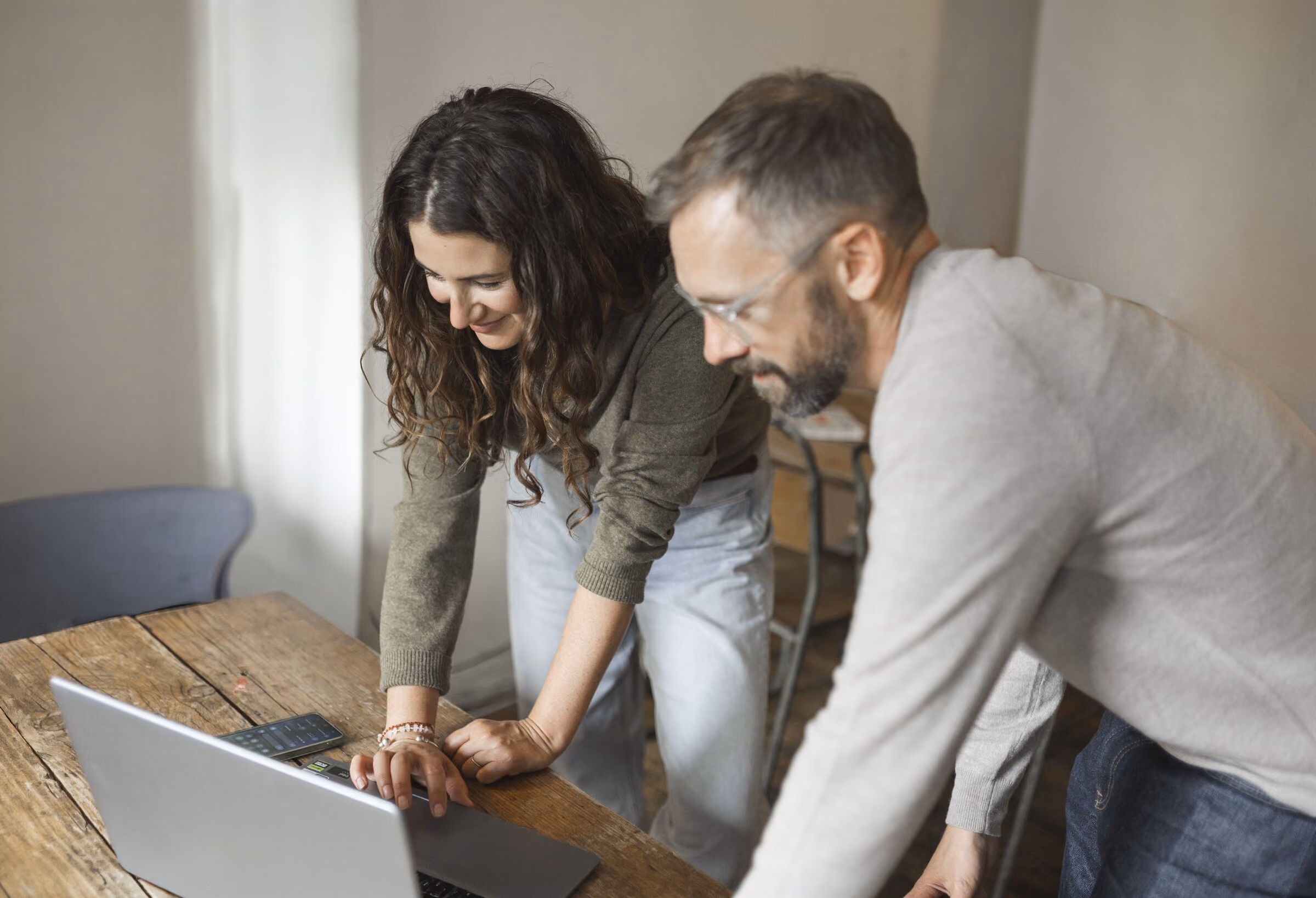 Two people leaning over a wooden table looking at a laptop screen, with a calculator also on the table.