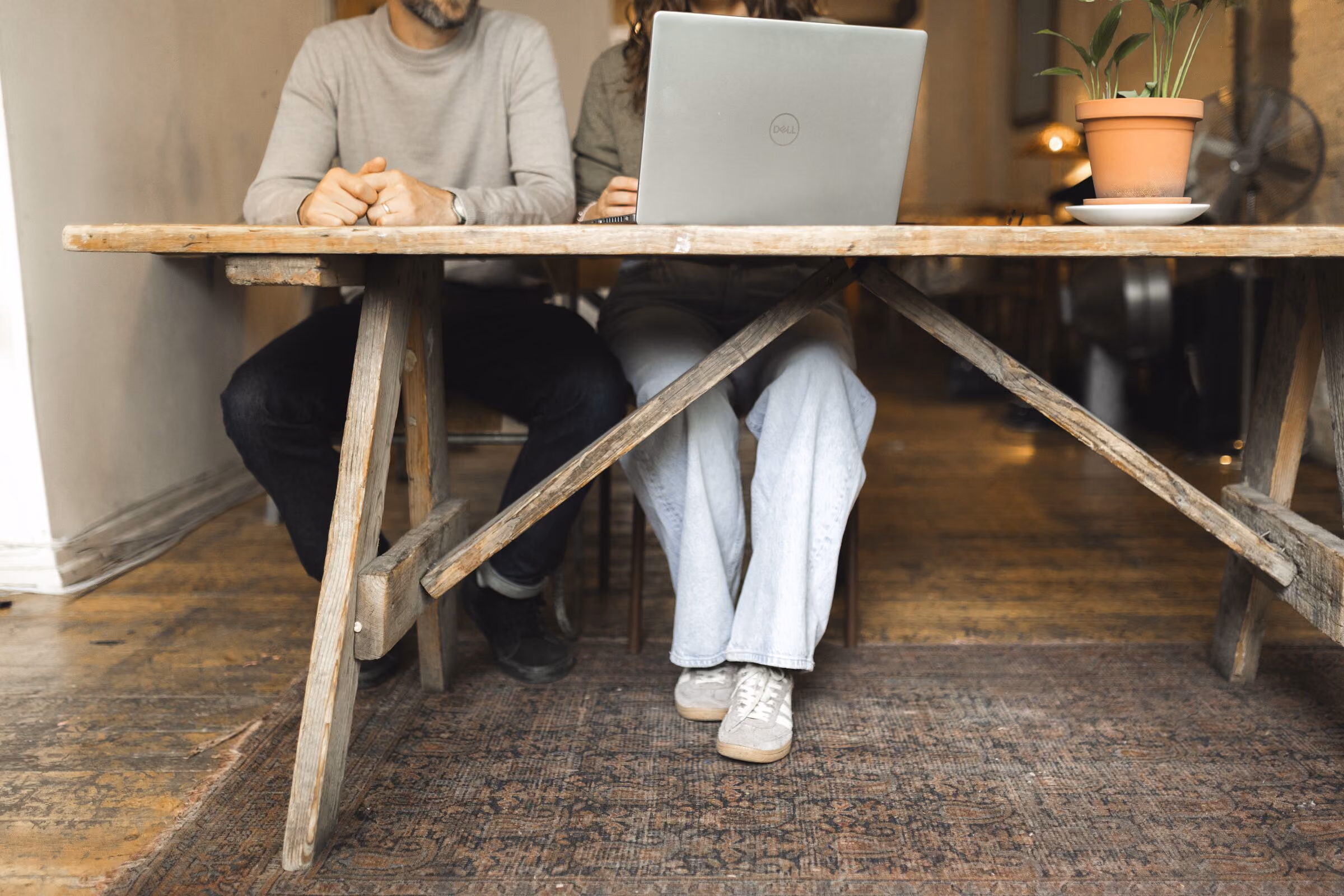 Two people sitting side by side at a rustic wooden table with a laptop and a potted plant on top, in a cozy indoor setting with wooden floors.
