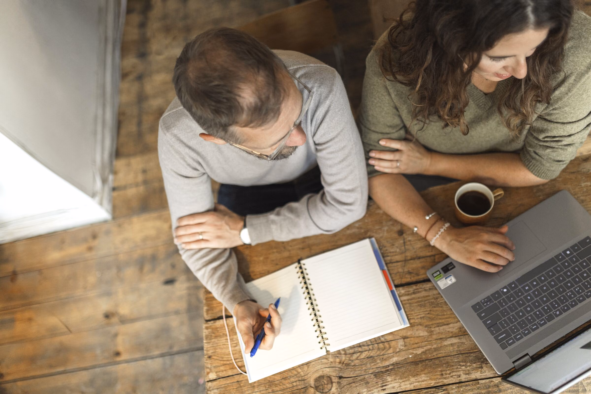 Top view of a man and woman sitting at a wooden table; the man writes in an open notebook with a blue pen while the woman uses a laptop and has a cup of coffee nearby.