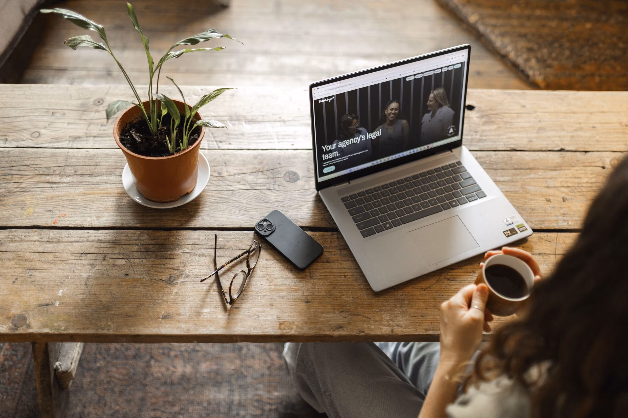 Person holding a cup of coffee sitting at a rustic wooden table with an open laptop displaying a legal agency website, a pair of eyeglasses, a smartphone, and a potted plant on the table.