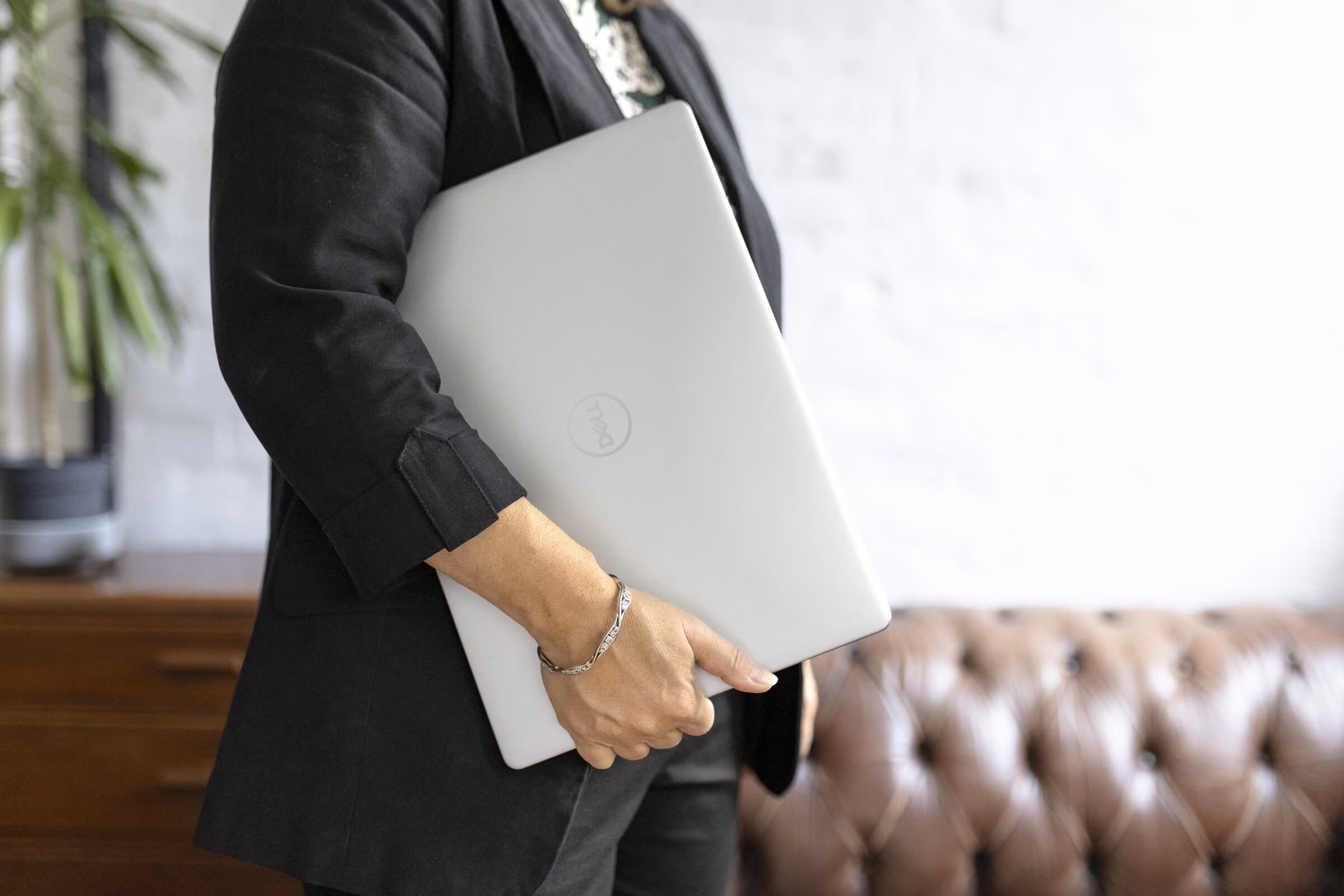 Person wearing a black blazer holding a closed silver Dell laptop against their chest, with a brown tufted leather couch and plant in the background.